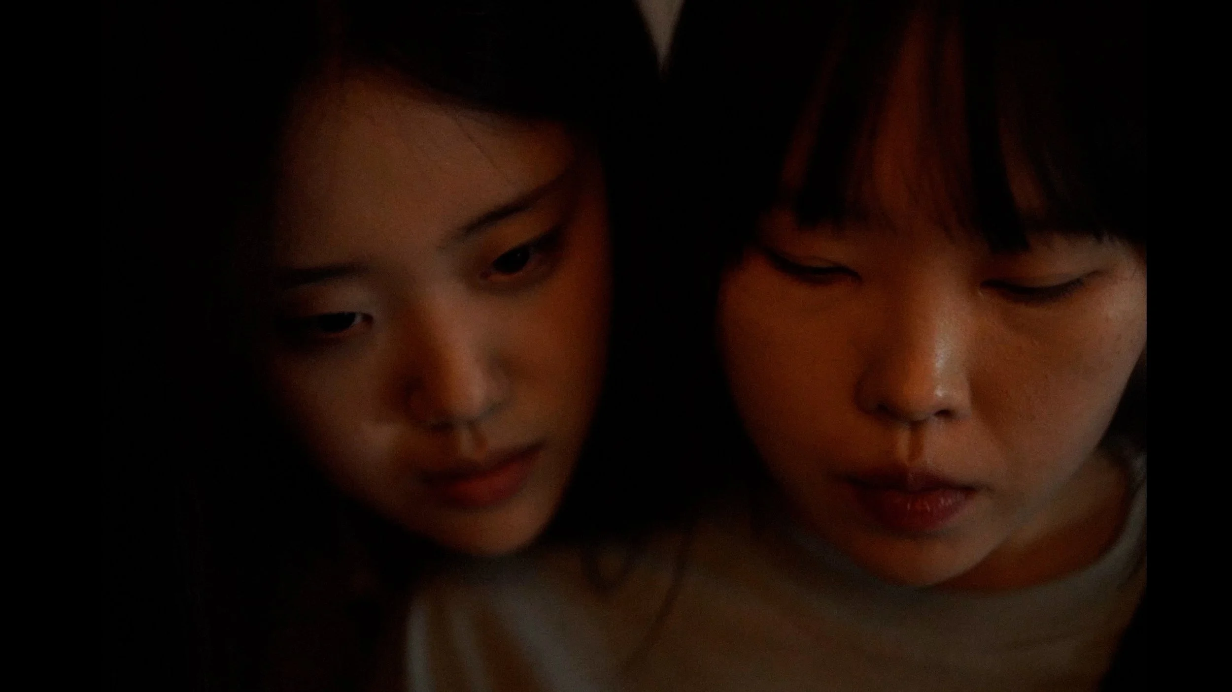 Close-up of two young women with dark hair and Asian features, one with a soft expression and the other with one eye open and the other eye closed, in a dimly lit environment.