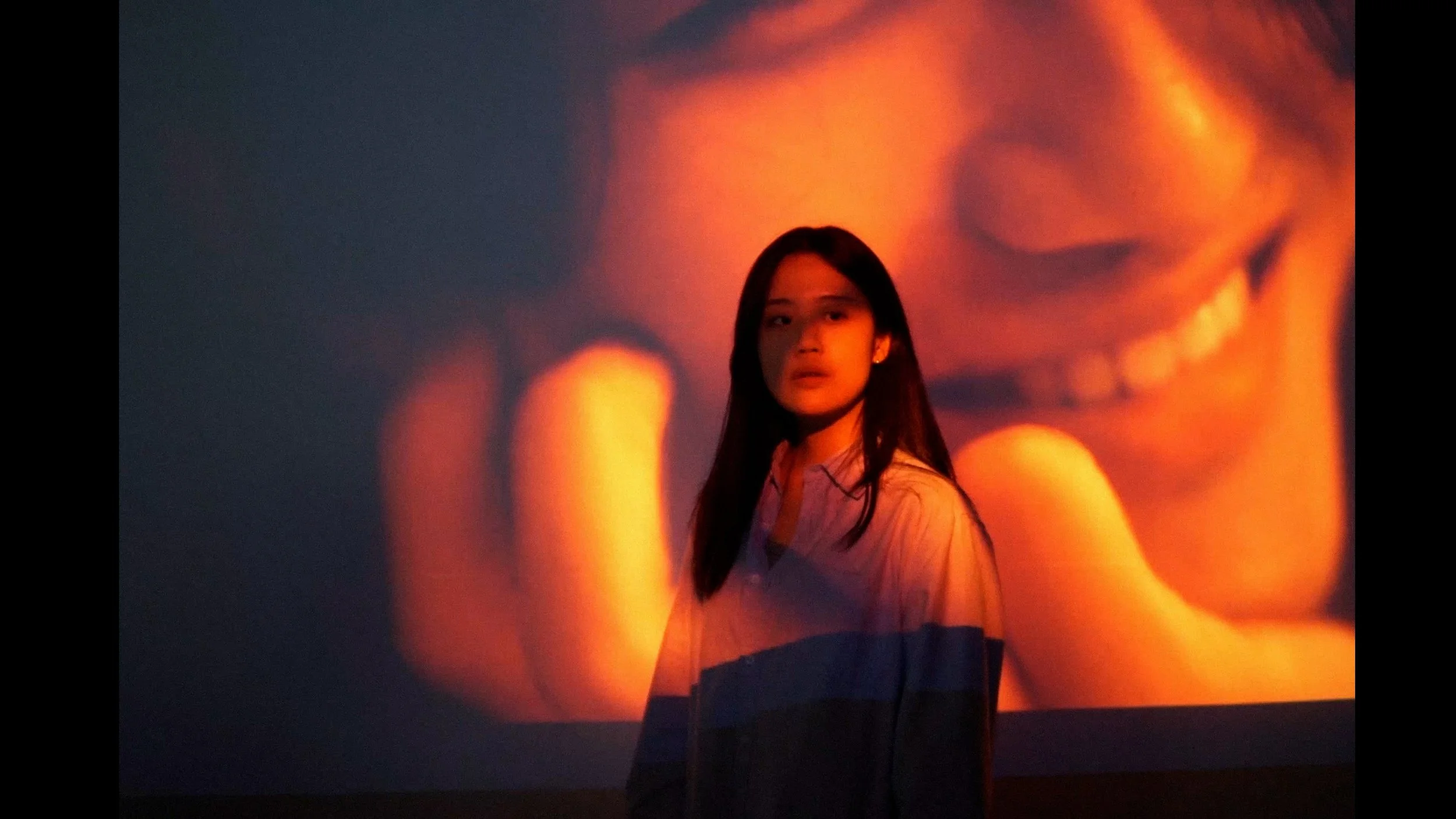 A young woman with long hair standing in front of a large projection or screen displaying a close-up of a smiling person's face in red and orange tones.