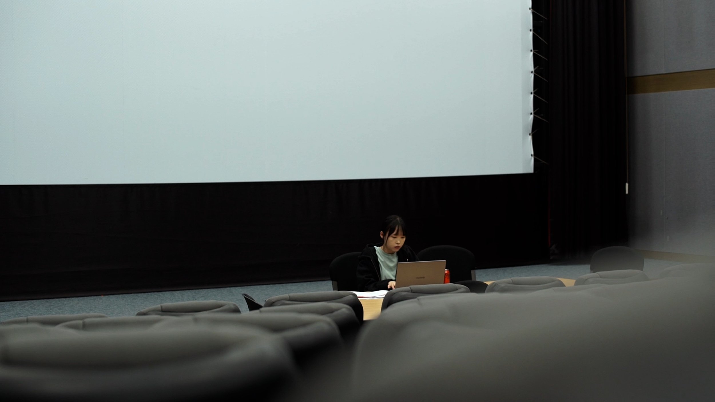 A young woman sitting at a table in an auditorium or theater, working on a laptop, with rows of empty seats and a large blank screen behind her.