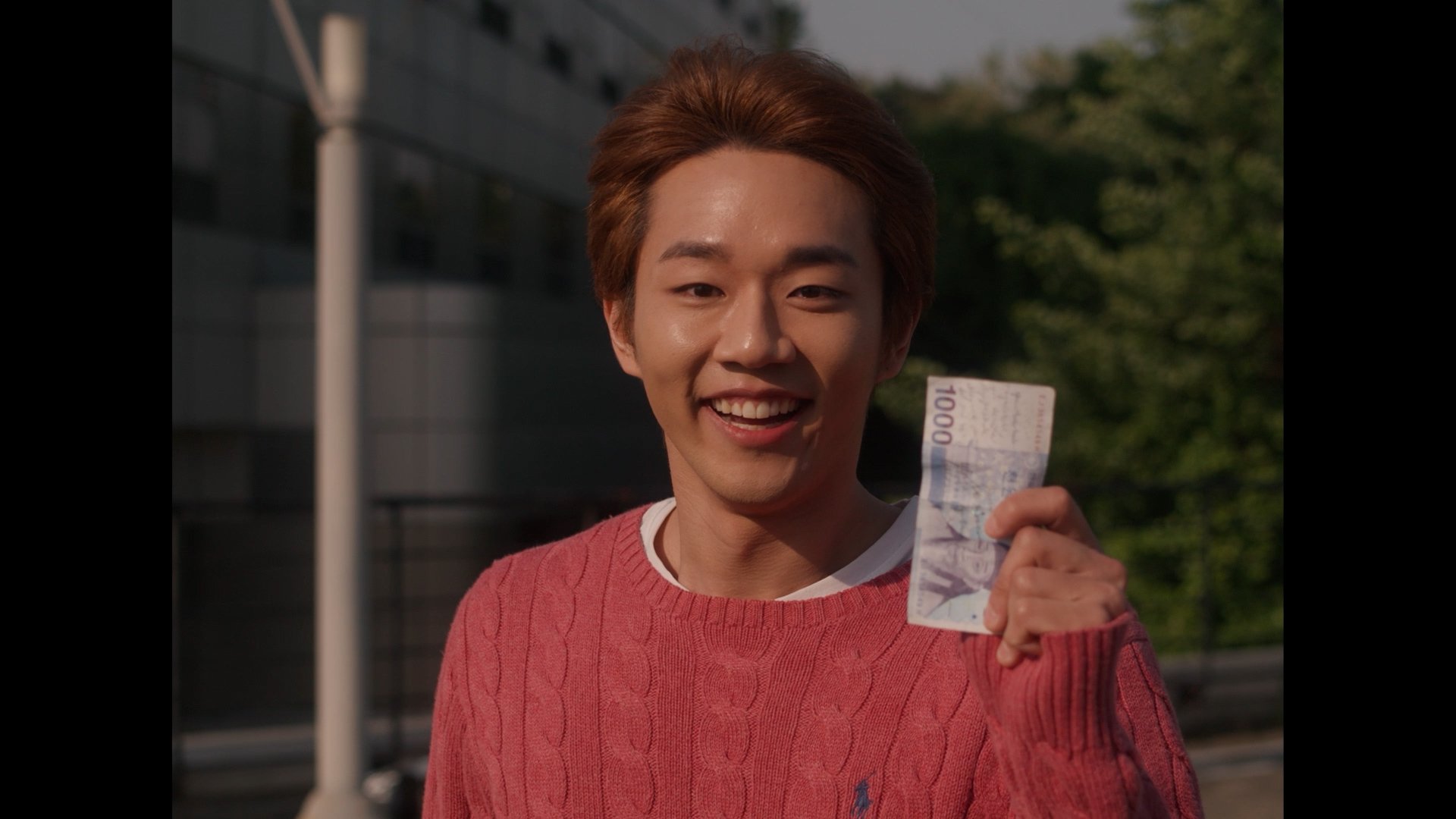 A young man with short brown hair, smiling and holding a 100-gram banknote, outdoors in front of a modern building and trees.