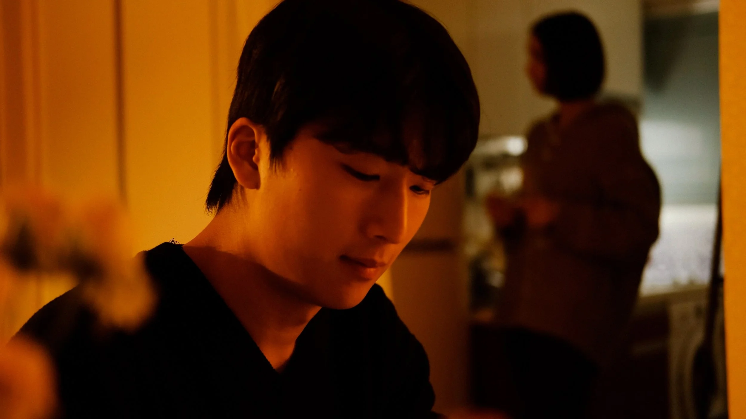 A young man with dark hair looking downward, captured in a warm, dimly lit room, with a woman blurred in the background.