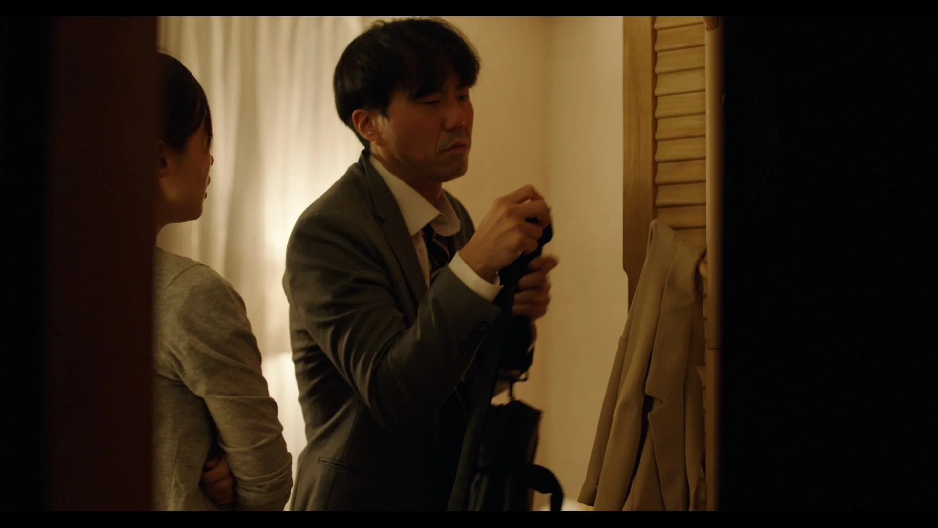 A man in a gray suit and tie is looking at a coat rack with jackets, with a woman in a light-colored shirt standing nearby in a warmly lit room.
