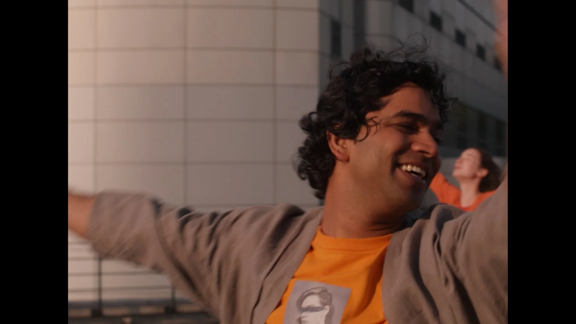 A man with curly dark hair smiling with his arms stretched out, wearing an orange shirt and gray jacket, in an urban environment with a tiled building wall behind him.