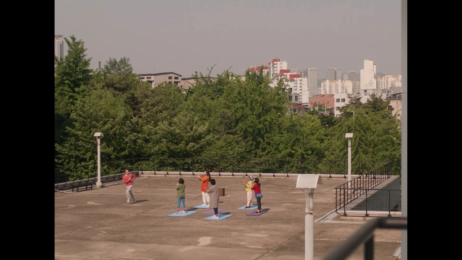 People practicing yoga outdoors on mats in an open area with trees and tall buildings in the background.
