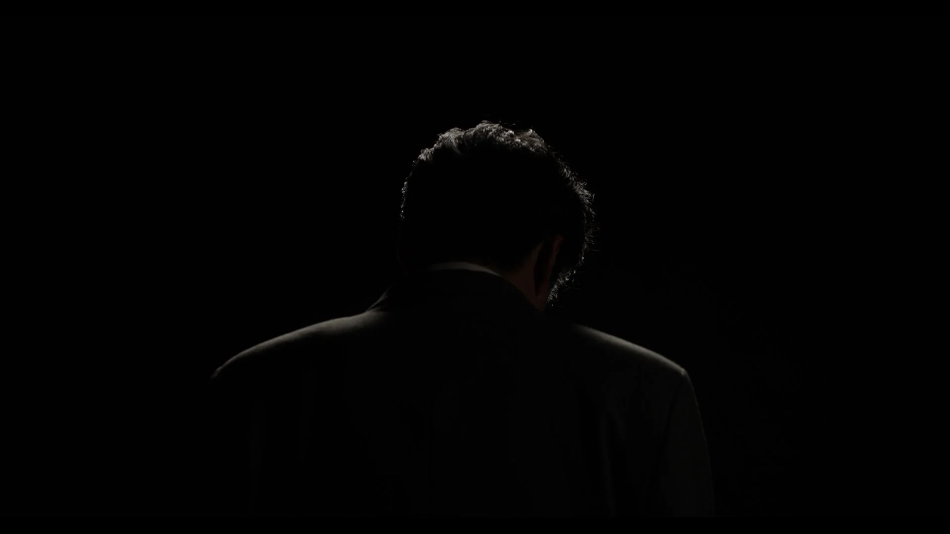Dark image of a man with curly hair seen from behind, wearing a suit, illuminated from the back and slightly above, with a black background.