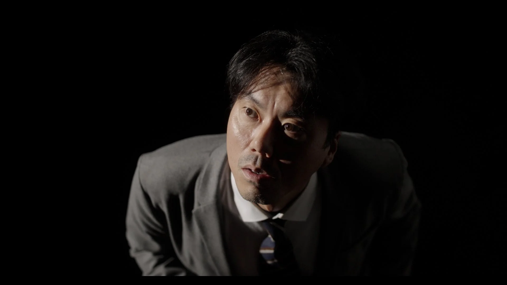 A man in a gray suit leaning forward with a serious expression, set against a black background.