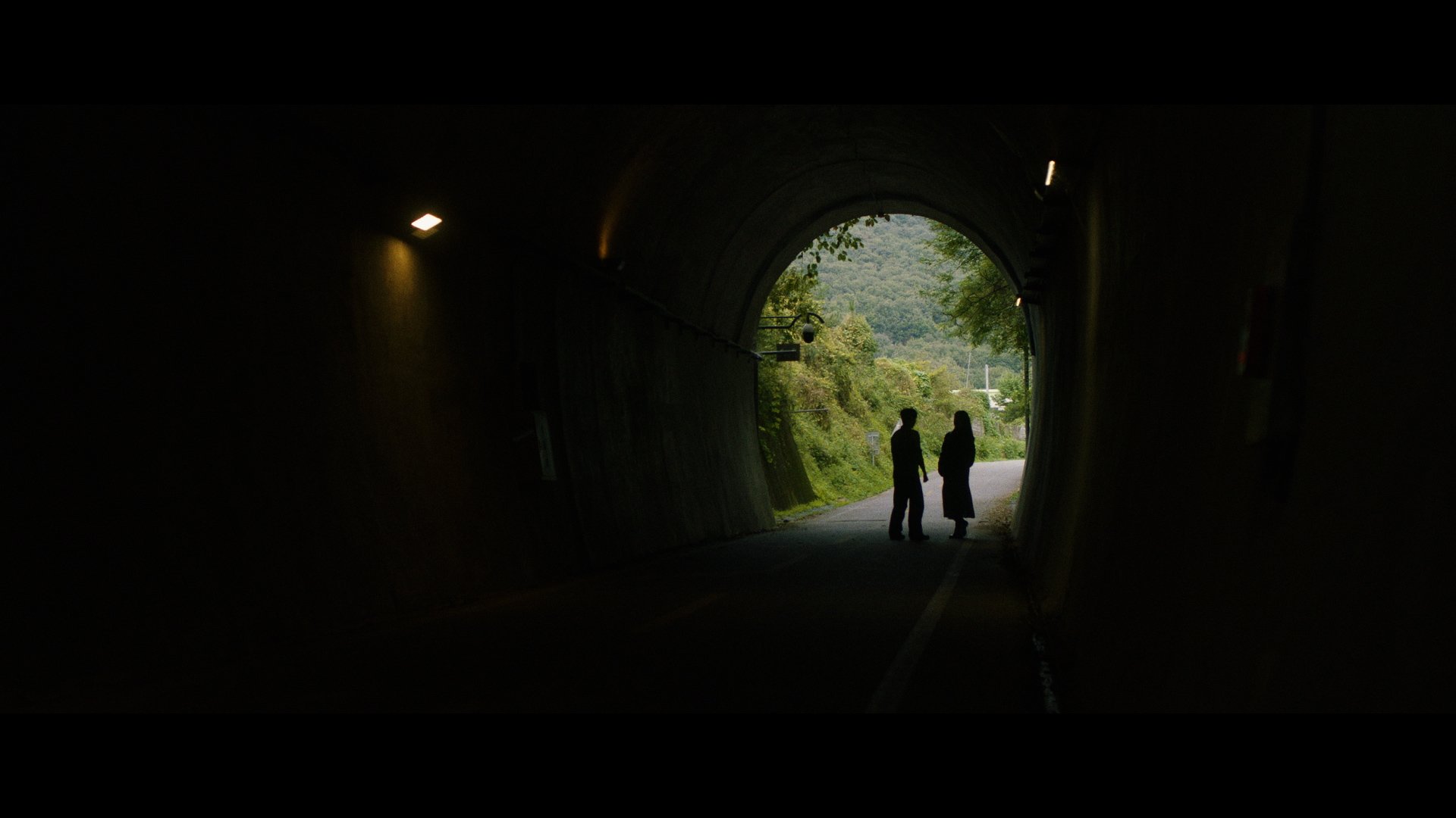 Two people standing at the entrance of a dark tunnel, with green trees and a landscape visible outside the tunnel.