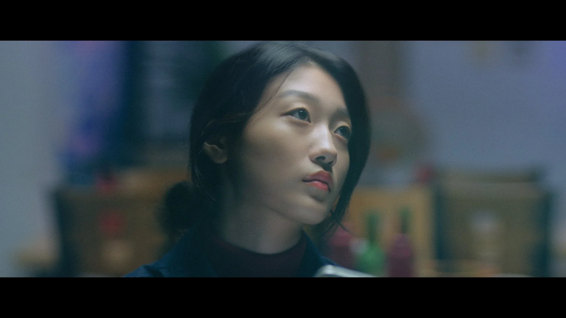 Close-up of a young woman with short black hair, wearing a black shirt, in a softly lit environment with blurred background, appearing thoughtful or pensive.