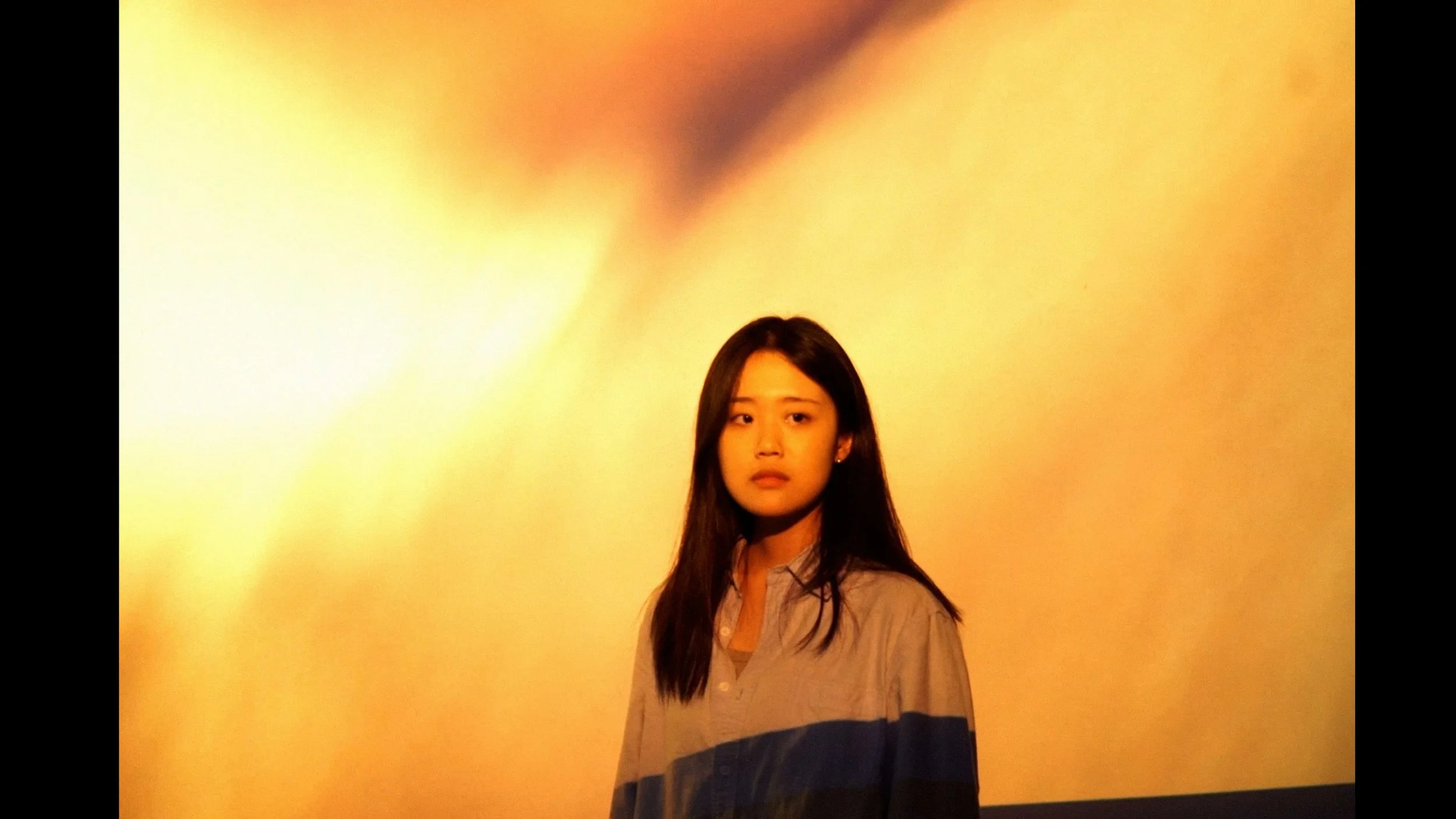 A young woman with long dark hair wearing a gray shirt standing in front of a plain yellow background.