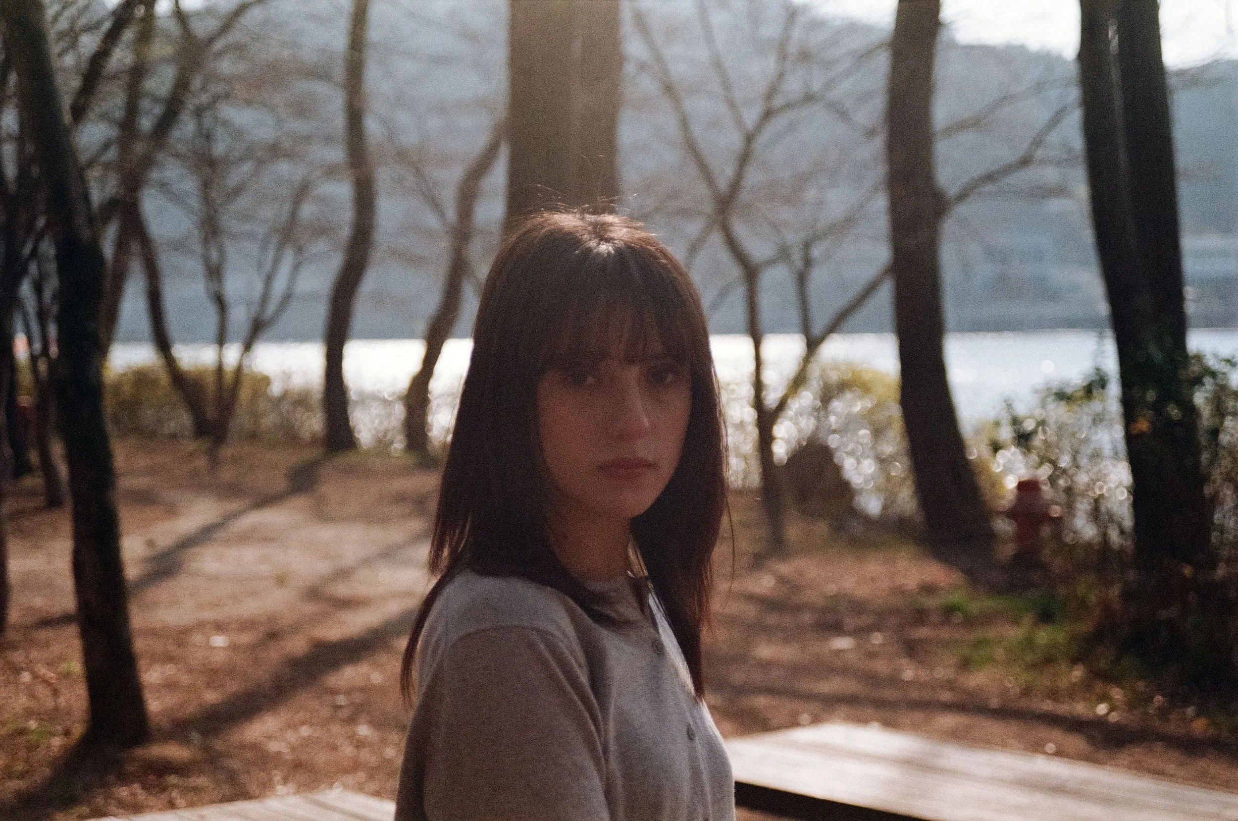A woman with medium-length dark hair and bangs standing outdoors in a park near a body of water, with trees in the background during daytime.