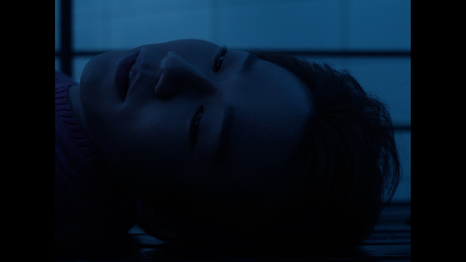 A person with dark hair lies on a surface, with a close-up view of their face, eyes closed, bathed in blue light.