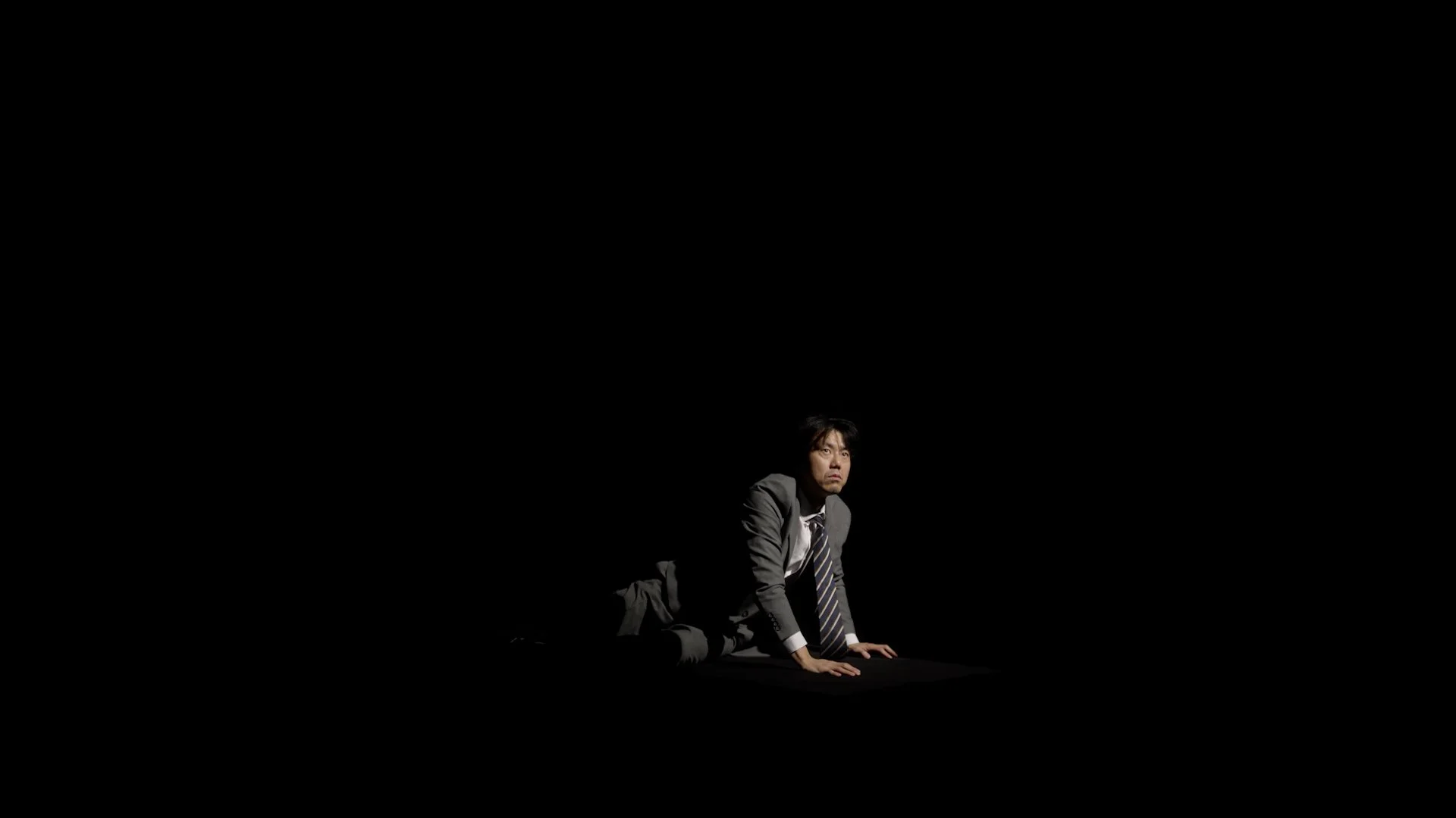 A man in a suit and tie is crawling on the ground in a dark environment, with minimal lighting highlighting his face and body.