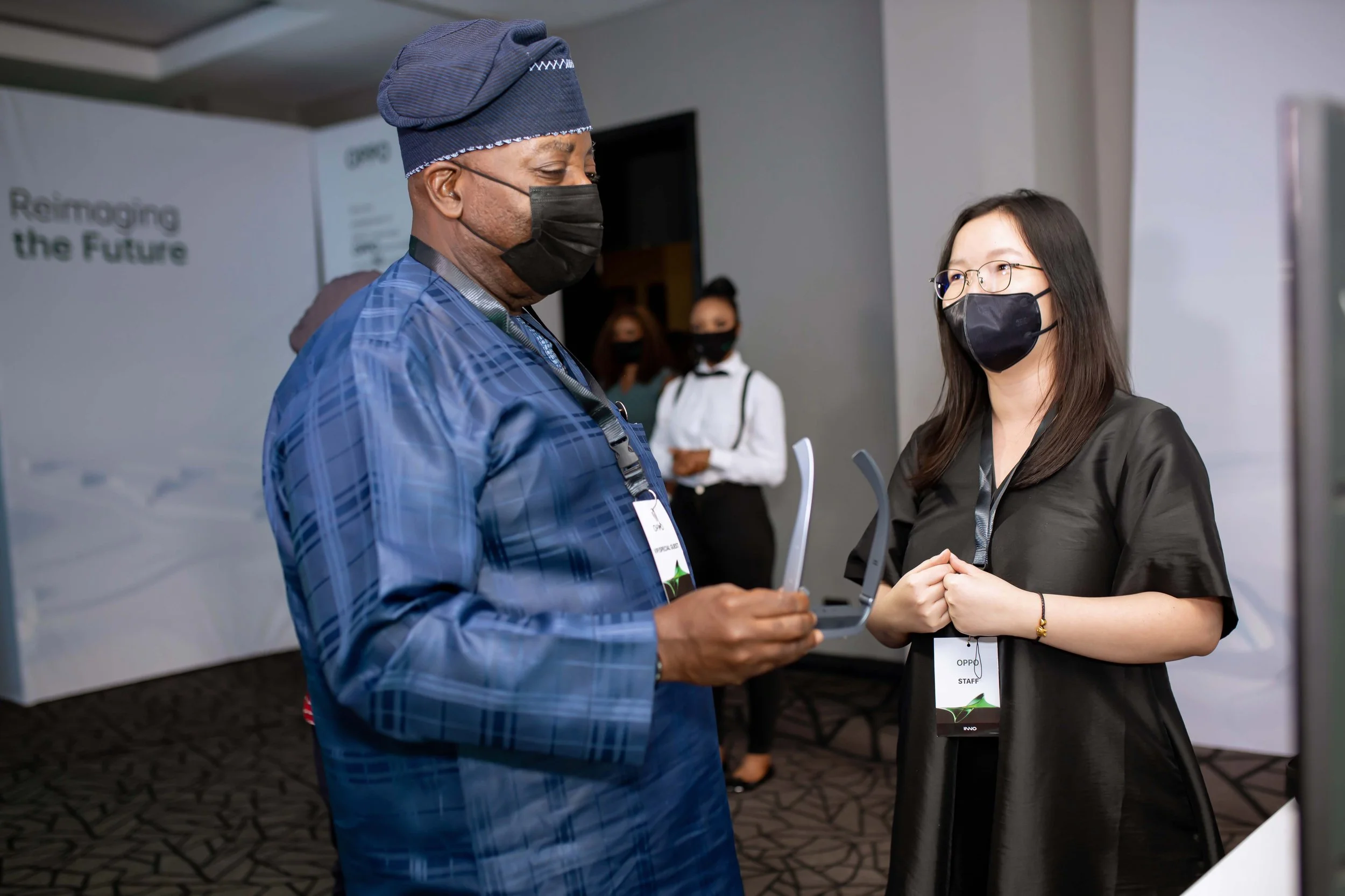 Two women wearing black masks and lanyards engaging in conversation at an indoor event. One woman has glasses and long hair, and the other wears a blue checkered shirt and a head covering.