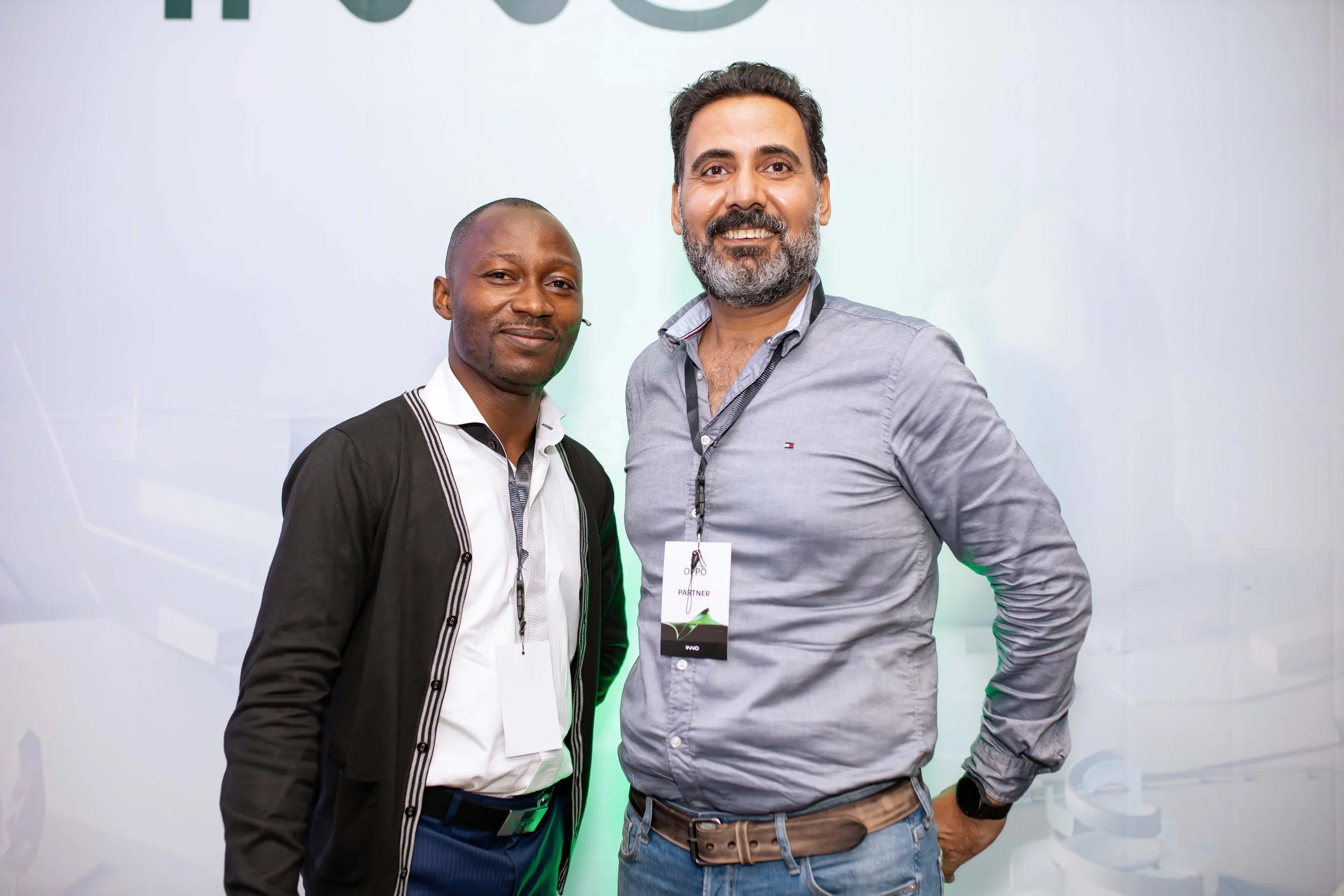 Two men standing together, smiling, at an event. One is wearing a white shirt with a black jacket, and the other is wearing a gray shirt with a lanyard and badge that says 'Partner'.