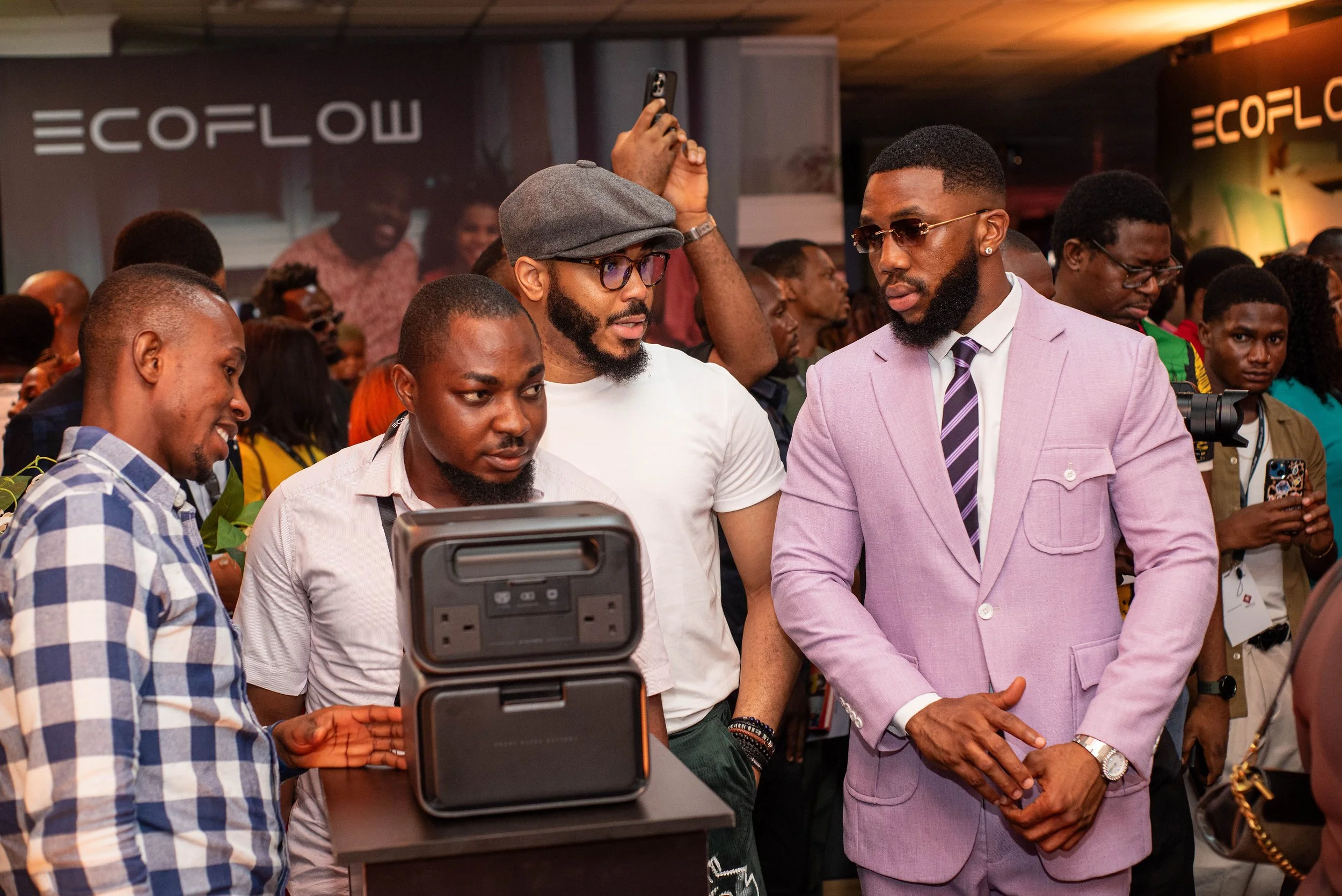 A man in a pink suit jacket and sunglasses is engaged in conversation with another man wearing a white shirt, glasses, and a gray cap, surrounded by a crowd of people at an indoor event.
