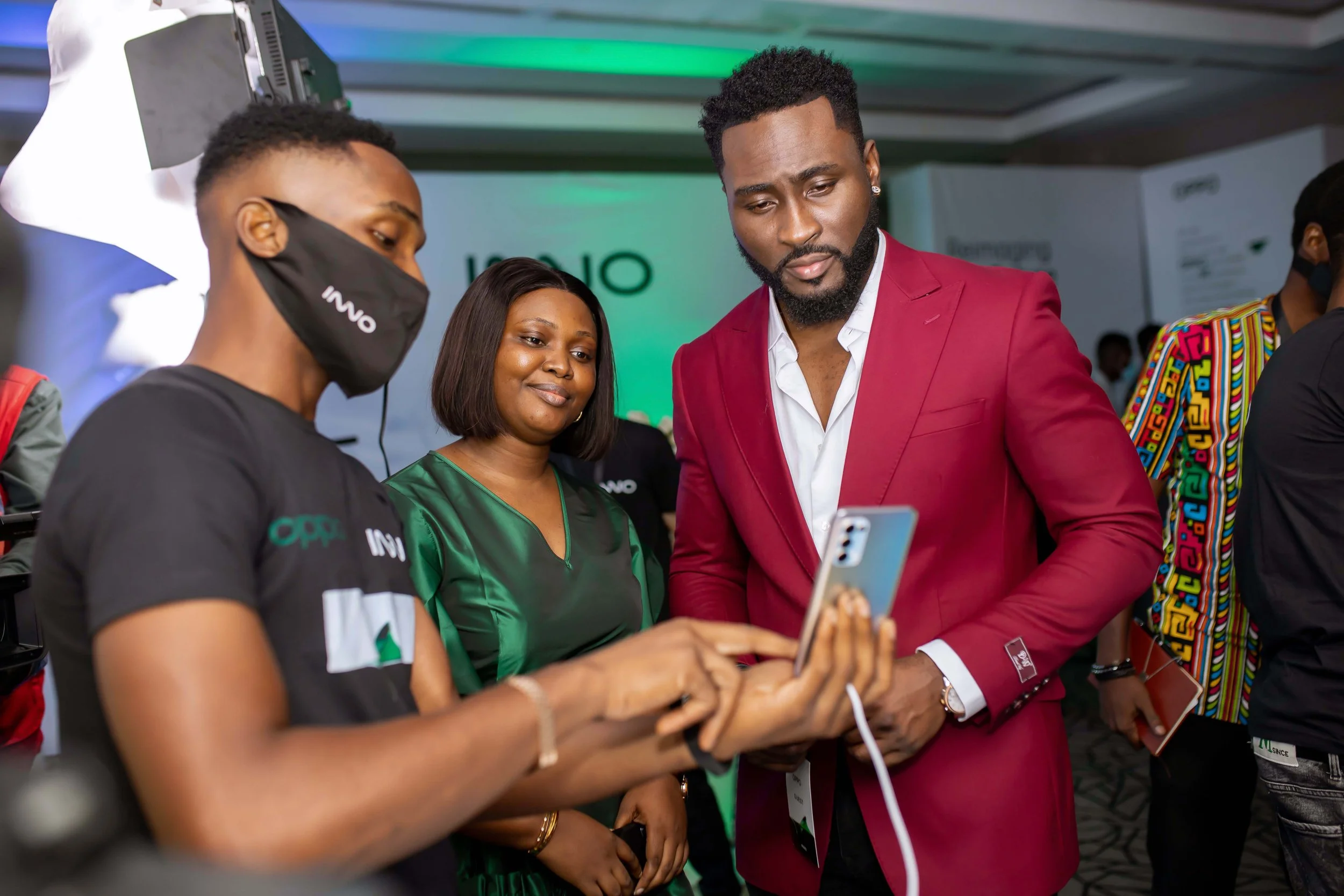 A group of people at an event, with a woman wearing a black face mask with the words 'INNO DAY' on it, a woman in a green shirt, and a man in a red suit holding a phone, looking at it together.