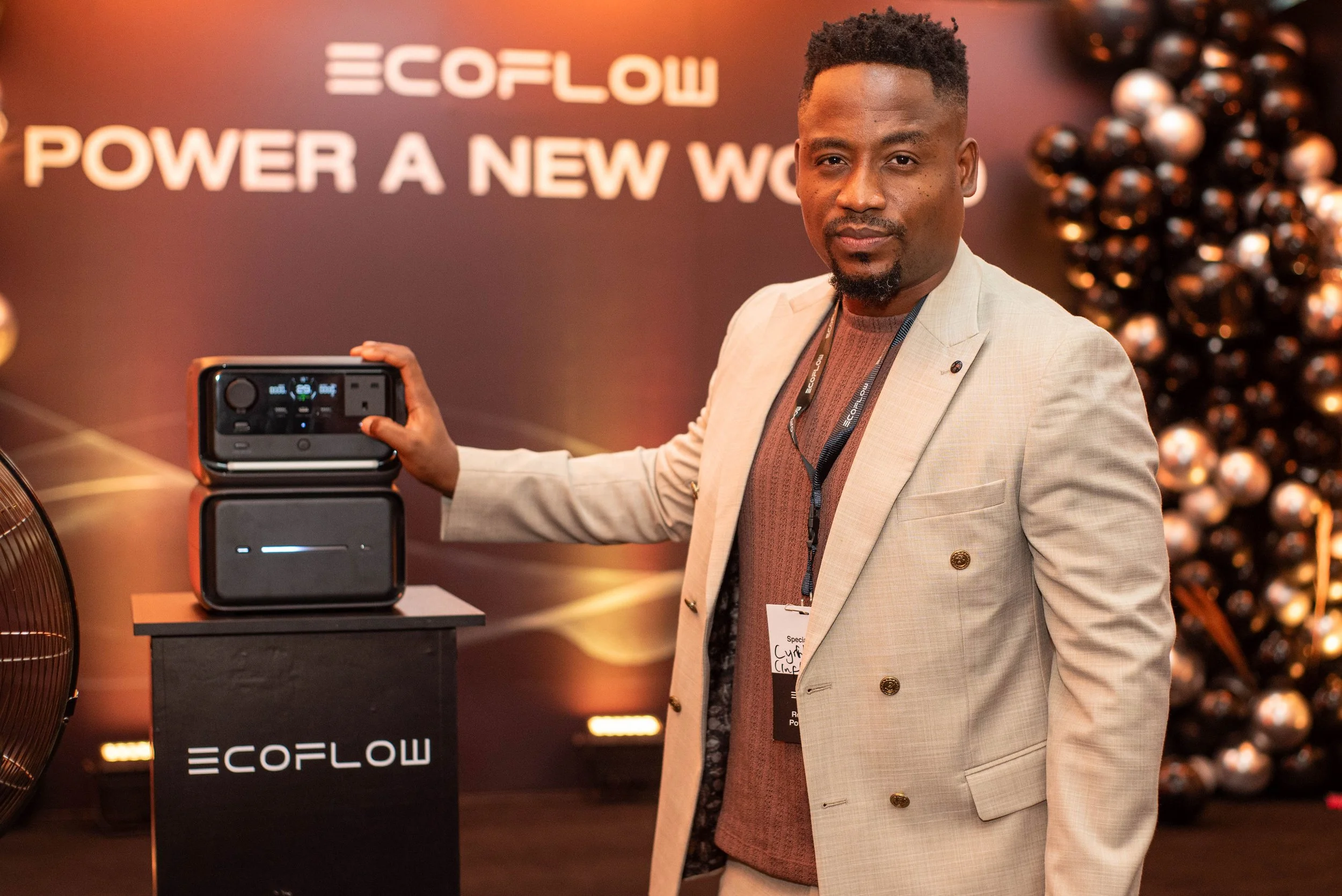 A man in a beige blazer standing in front of a display with the logo and name 'ECOFLOW' and the text 'POWER A NEW WORLD'. He is holding a portable power station.