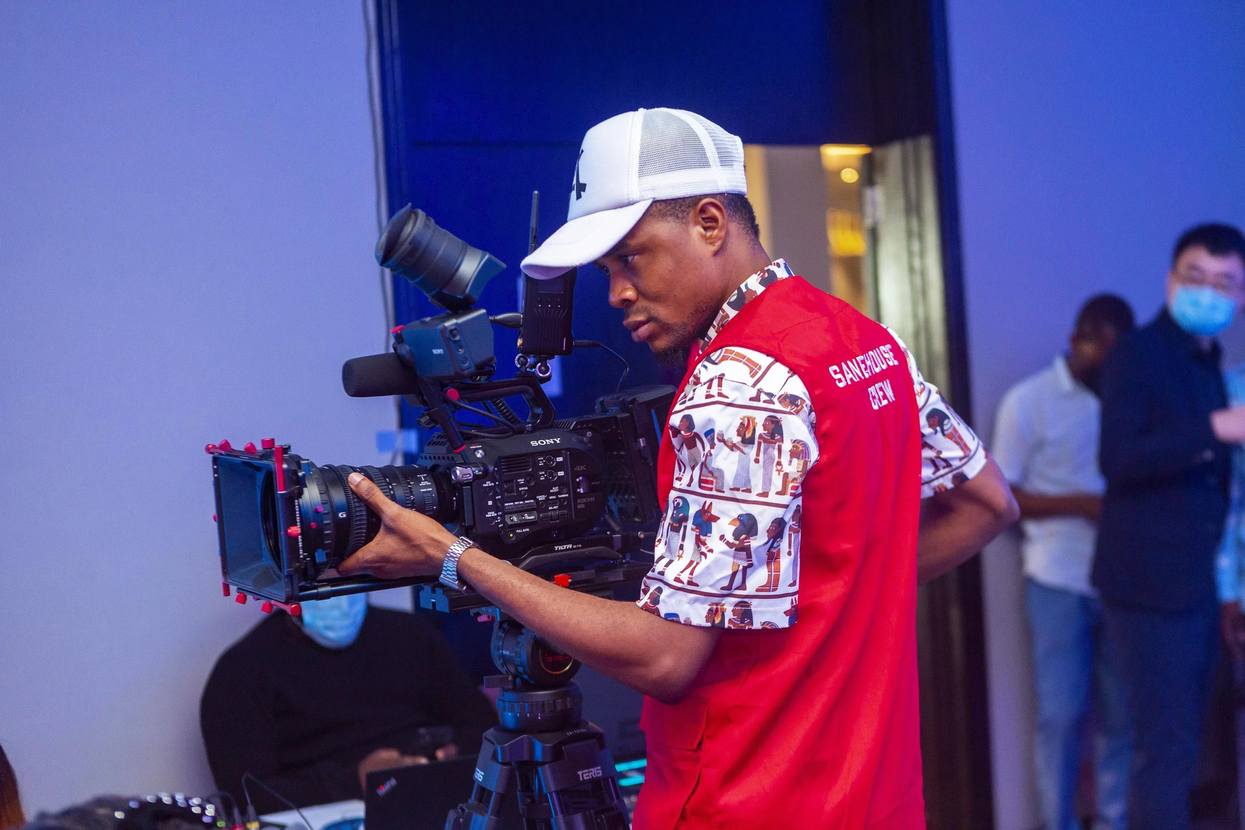 A man operating a professional video camera at an indoor event, wearing a white cap and a colorful patterned shirt. In the background, people, including a man with a face mask, are visible.