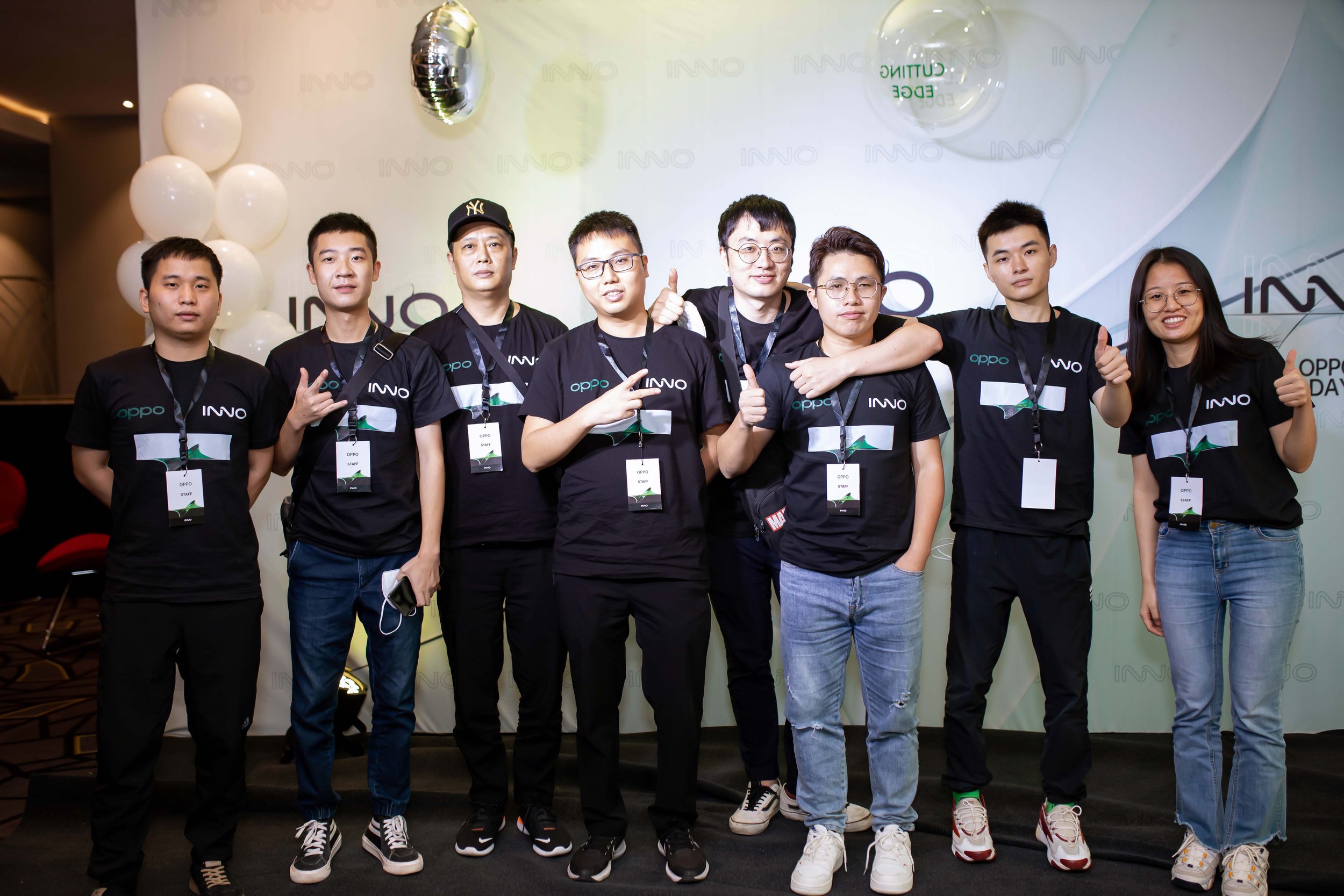 Group of eight young adults standing in front of a backdrop with balloons, wearing black shirts with the Oppo logo, some making hand gestures, giving thumbs up, and smiling.