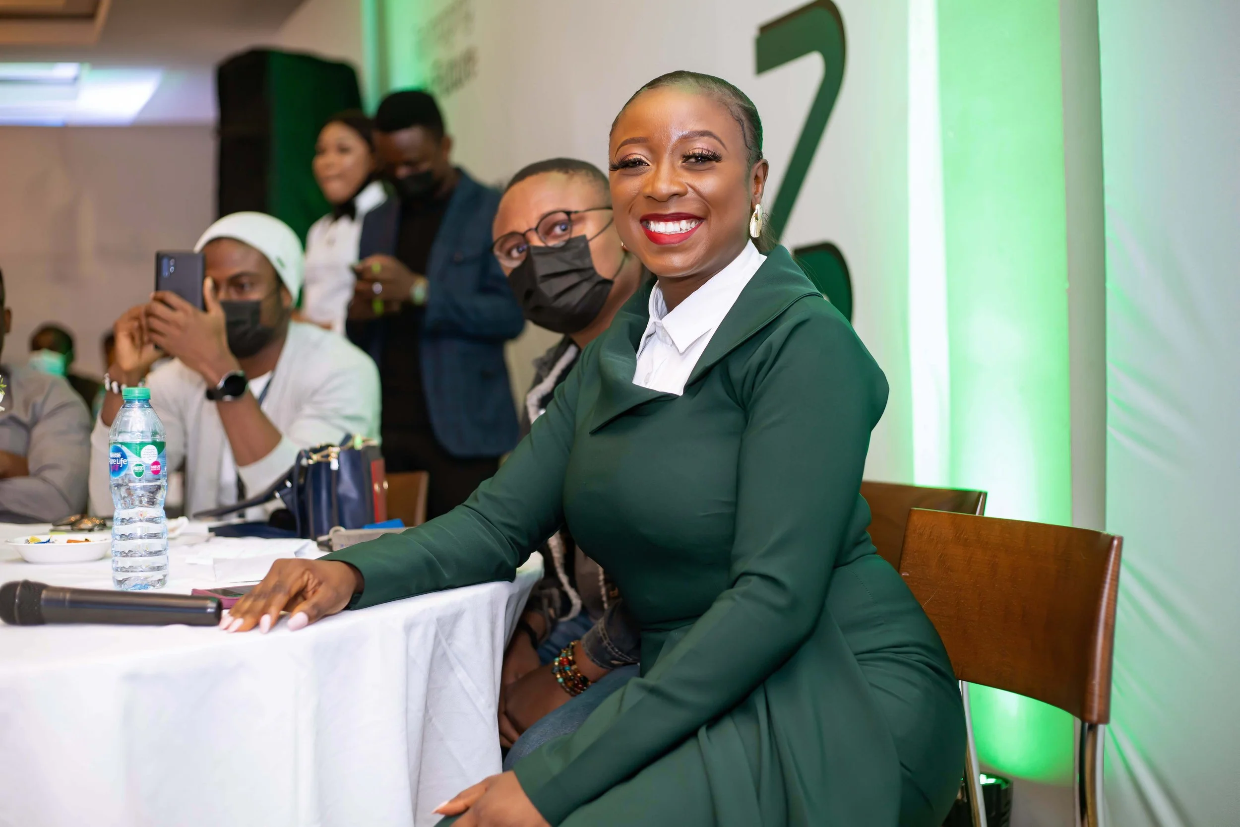 A smiling woman in a dark green suit sitting at a conference table, surrounded by others at a professional event. The background features a large green and white logo on the wall.