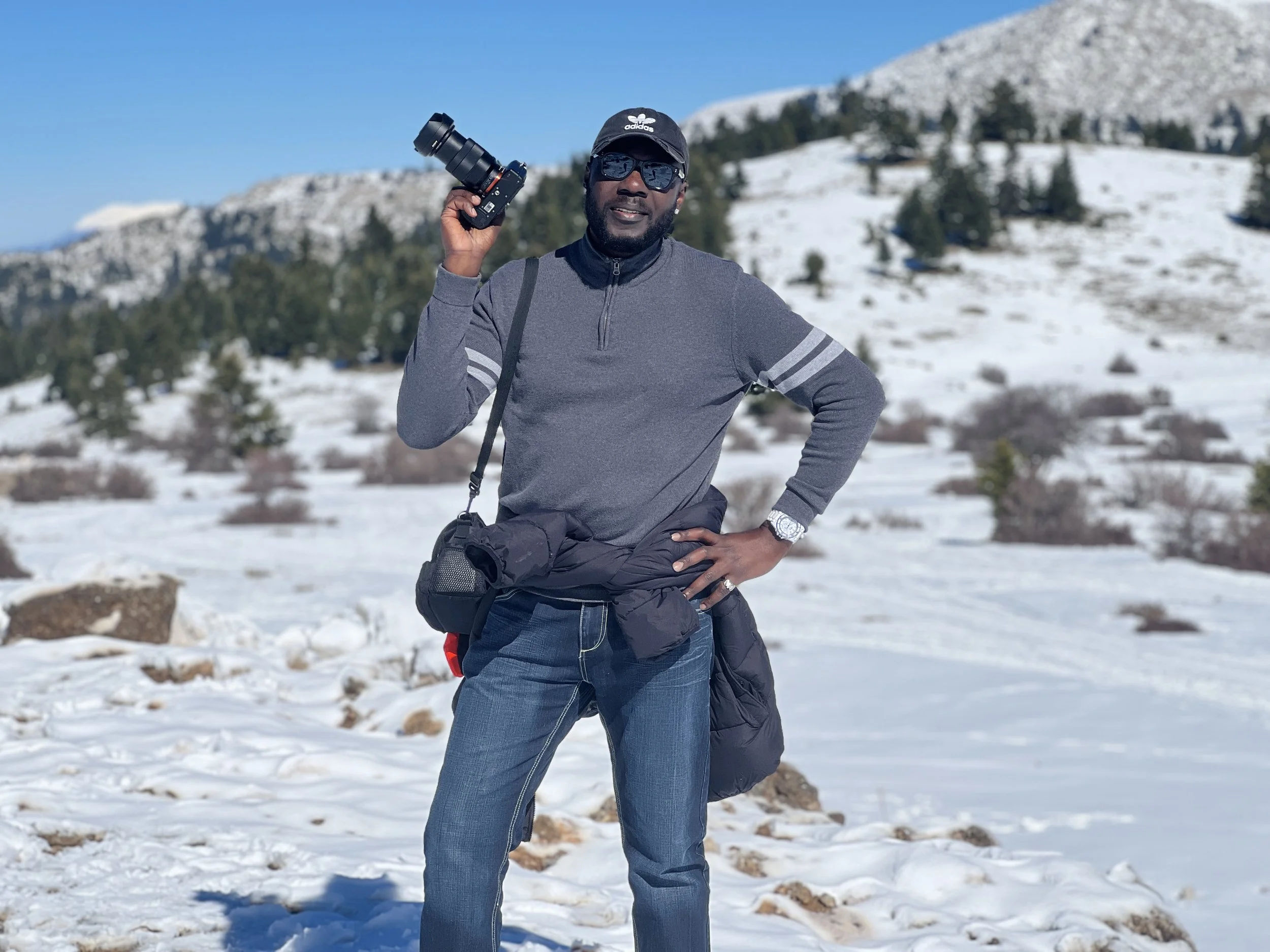 A man wearing a dark cap, sunglasses, grey quarter-zip sweater, and blue jeans stands in a snowy mountain landscape holding a camera upright in his right hand.