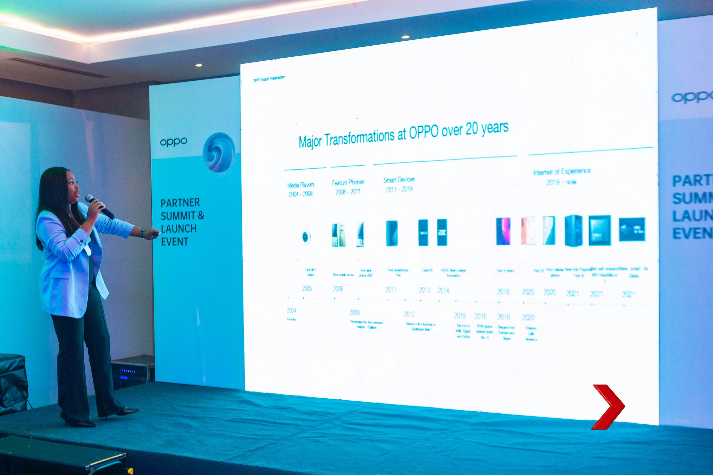 A woman giving a presentation at a conference, standing on a stage beside a large screen displaying a slide titled 'Major Transformations at OPPO over 20 years'. The slide highlights different phases in the company's history with illustrations of OPPO smartphones at the bottom.