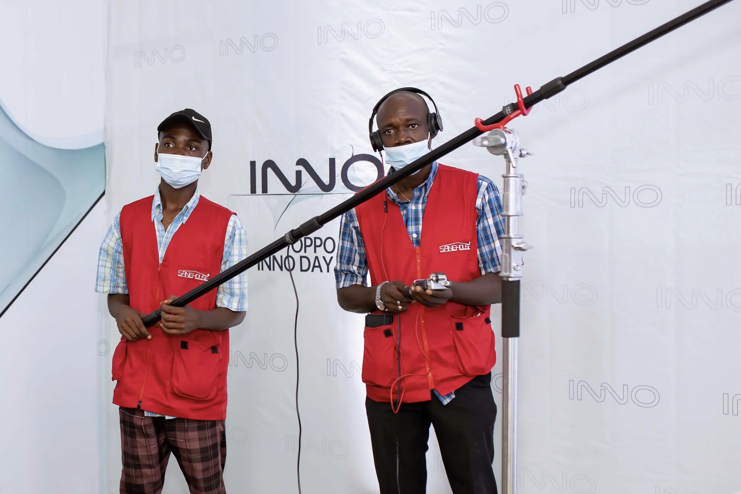 Two Sane House Media  crew members standing behind professional filming equipment at an event. One man is holding a control device, and the other is observing with a headset. The backdrop has the word INNO repeated across it.