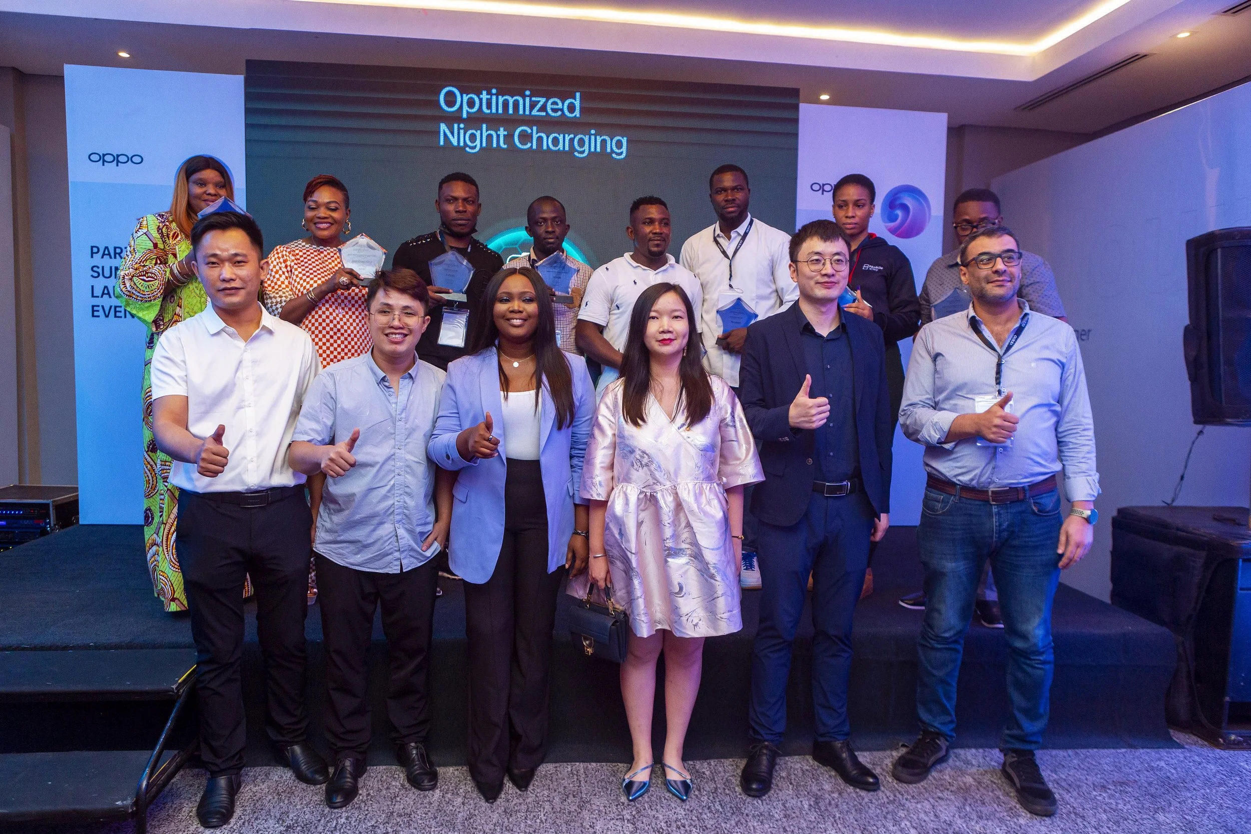 OPPO staff and guests standing together on a stage at an event. Some are holding awards, and a large screen behind them displays 'Optimized Night Charging.'