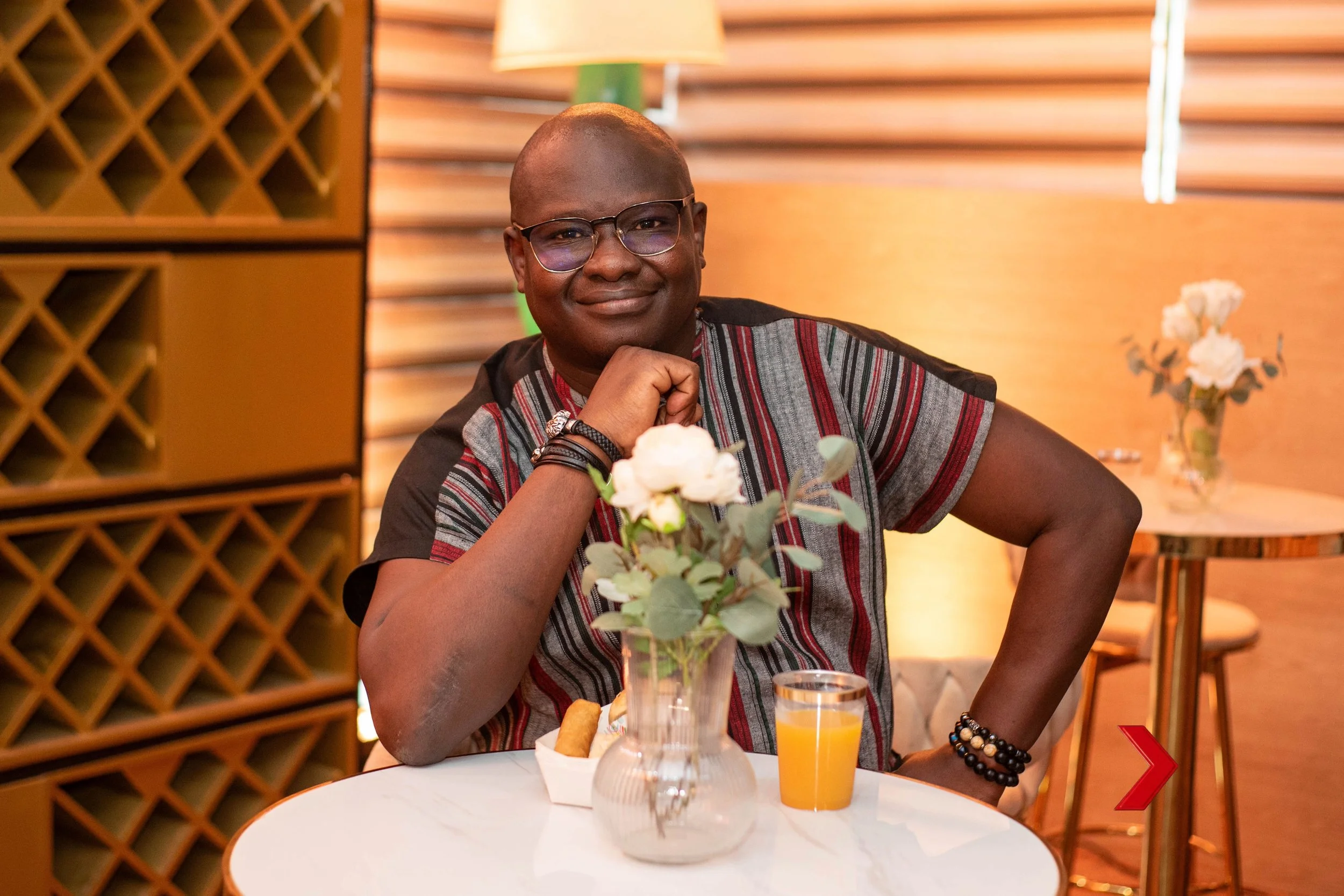 A man with glasses sitting at a table in a restaurant or cafe, smiling with his chin resting on his hand. The table has a vase with white flowers, a glass of orange juice, and a small plate of food. The background features wooden décor and warm lighting.