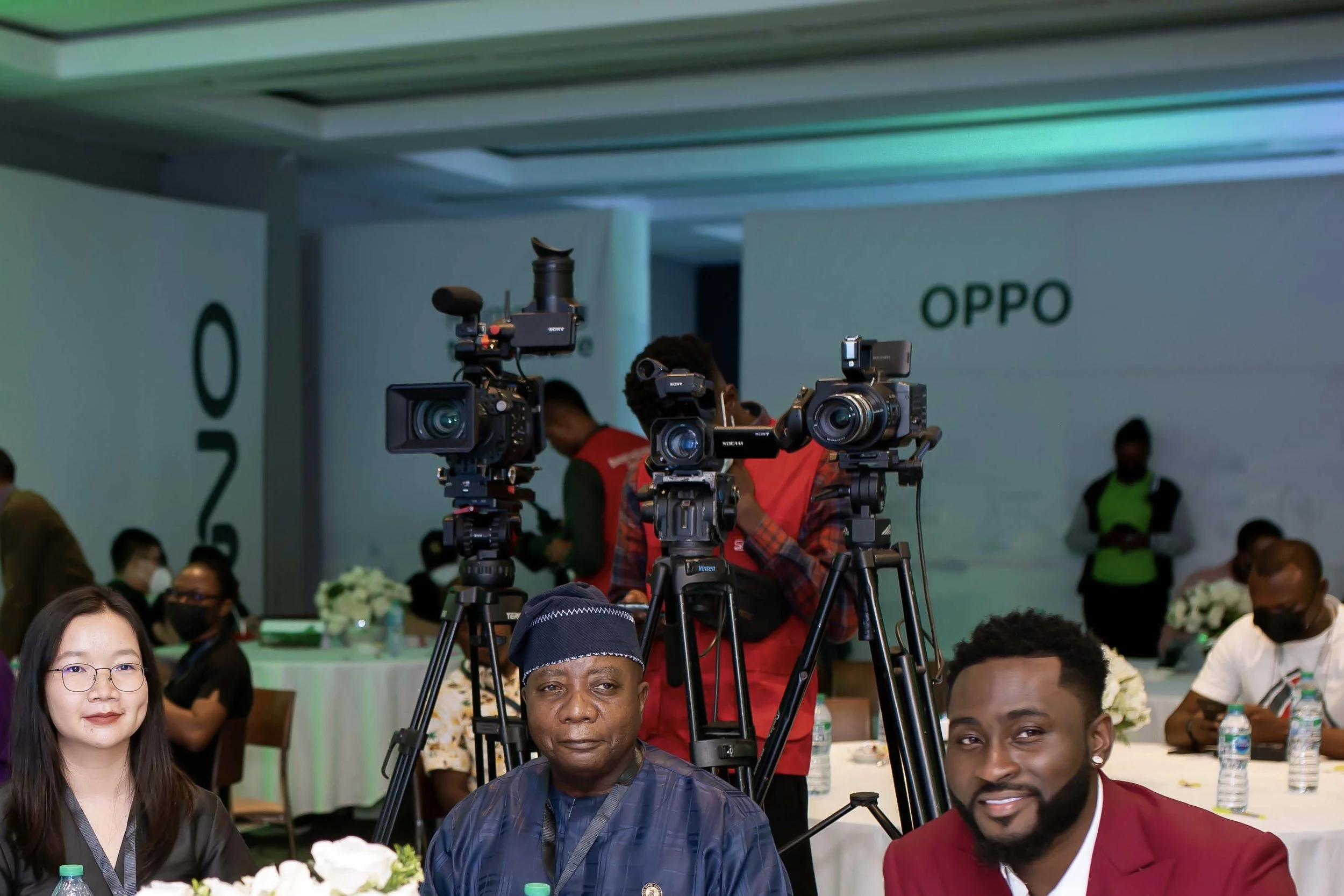 Group of people at an event with professional video cameras set up for recording media coverage, with some attendees seated at tables and others standing, in a large indoor venue with white walls and a green and white color scheme.