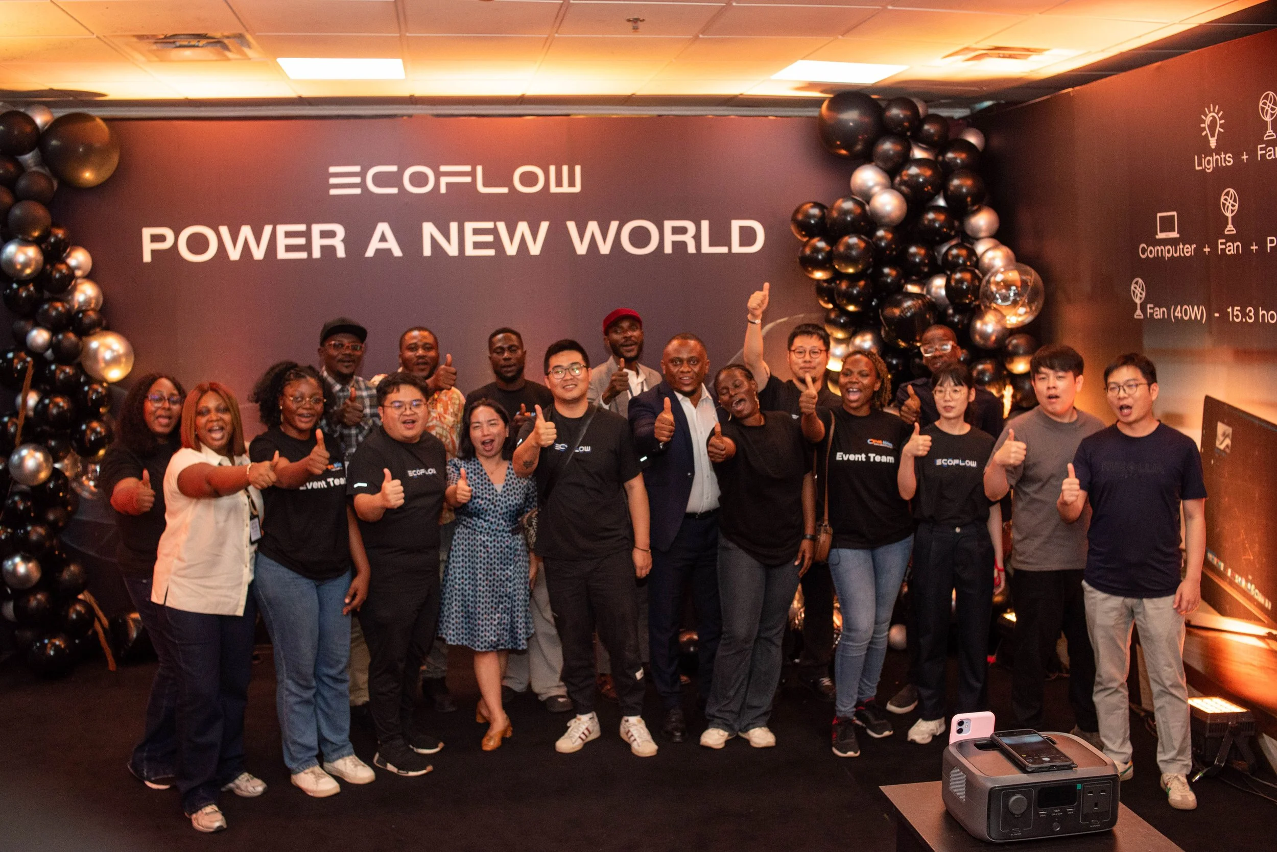 Group of diverse people smiling, giving thumbs up, and posing for a photo in front of a large dark purple wall with balloons and the words 'EcoFlow Power a New World' written on it, at an event or celebration.