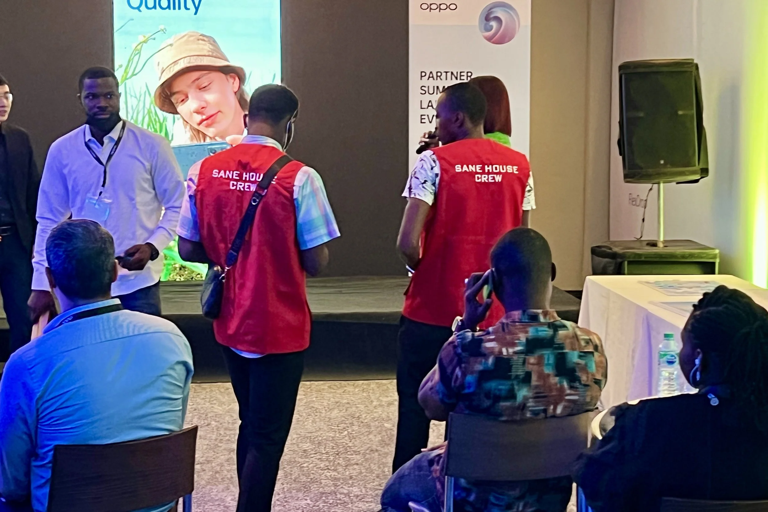 People attending a conference or event, seated and standing around, with some wearing red vests labeled 'SANE HOUSE CREW'. A large screen displays a woman's face with the words 'Quality' in the background, and there is a speaker on the right side.