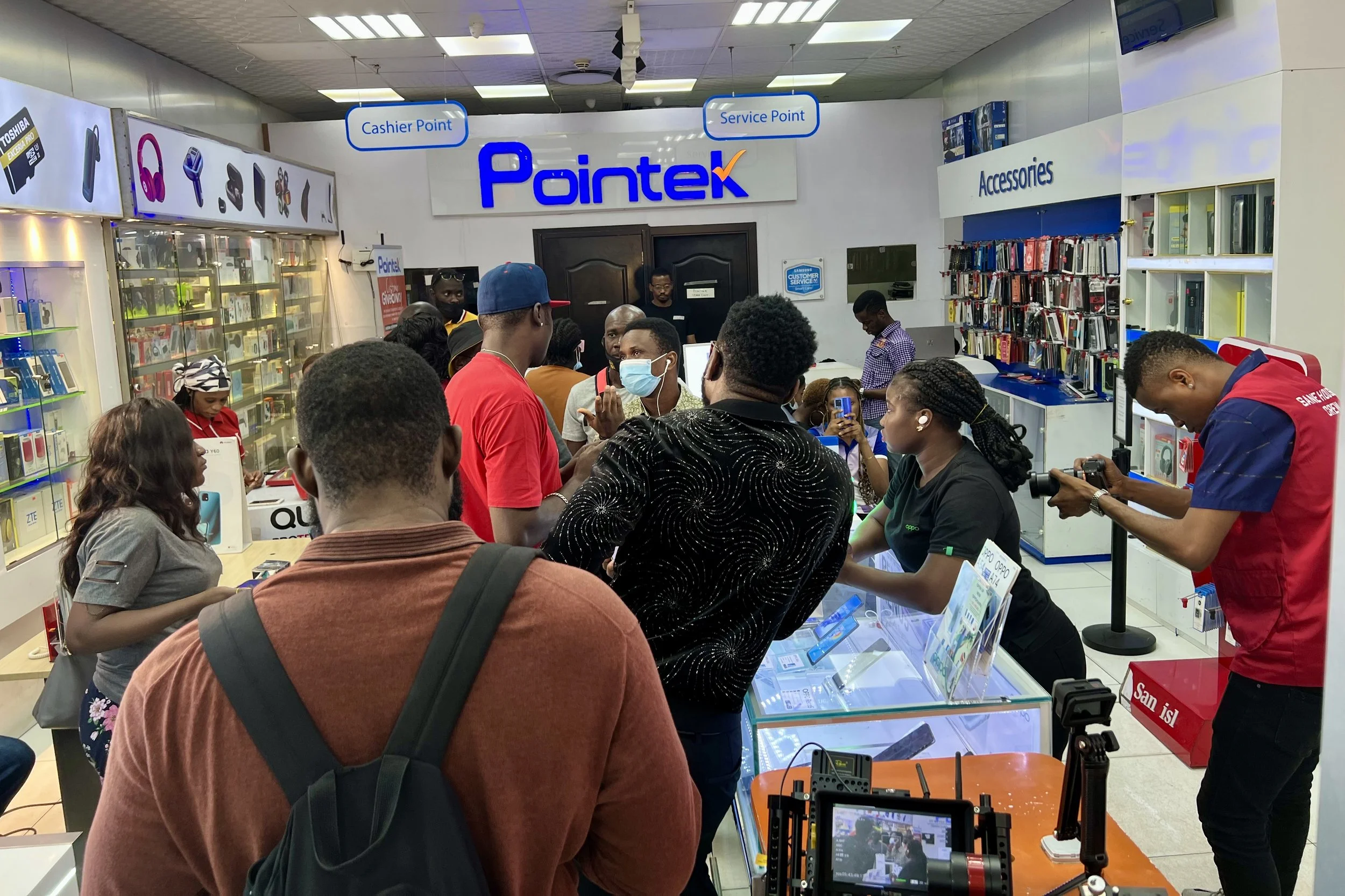Sane House Media director speaking to cast and crew during a video shoot at a tech store, with phone and accessory displays in the background.