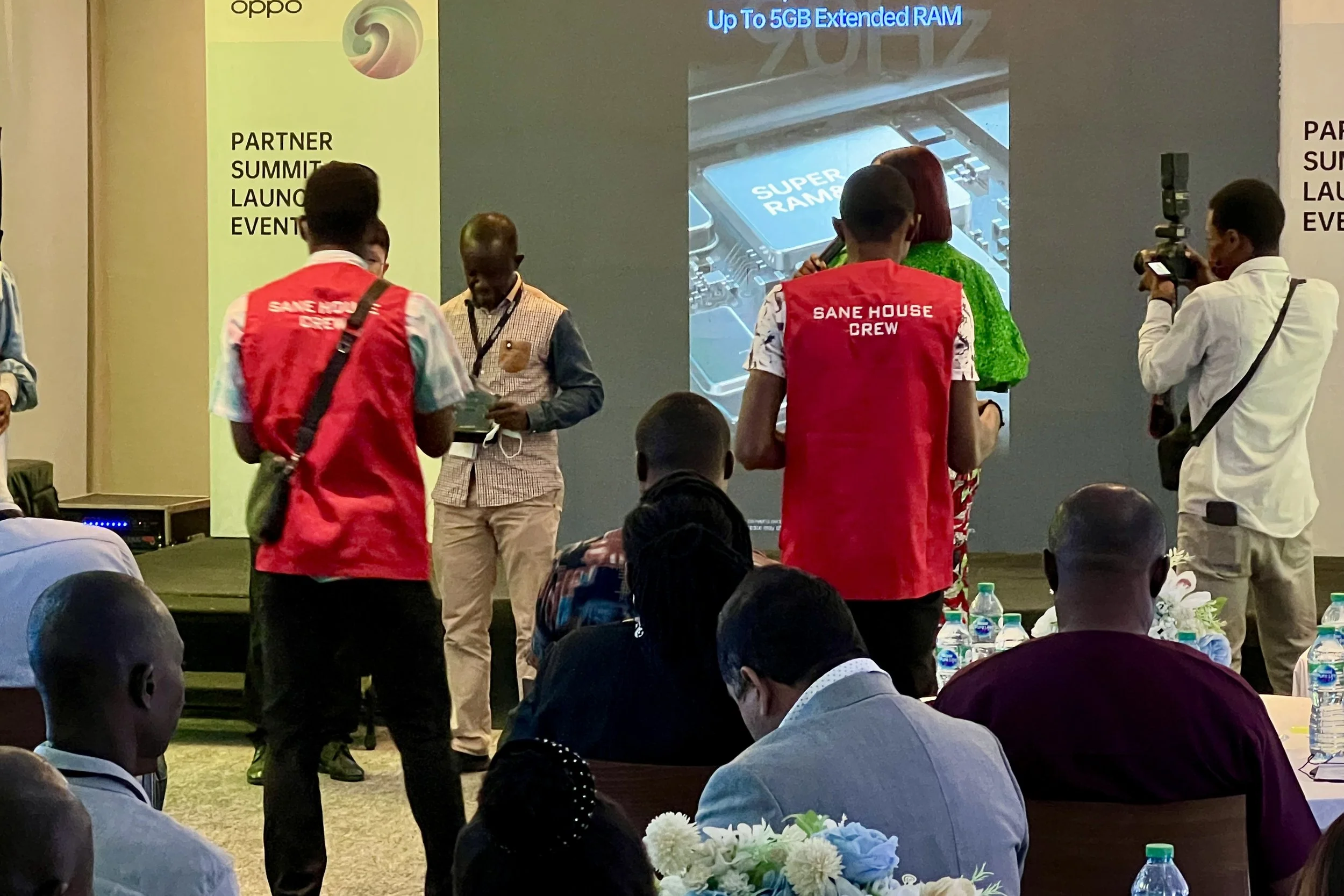 At a conference or summit, people are gathered in front of a large presentation screen, which displays information about RAM and a tech device. Some attendees are taking photos or talking, and several have red vests labeled 'SANE HOUSE CREW.' The roo