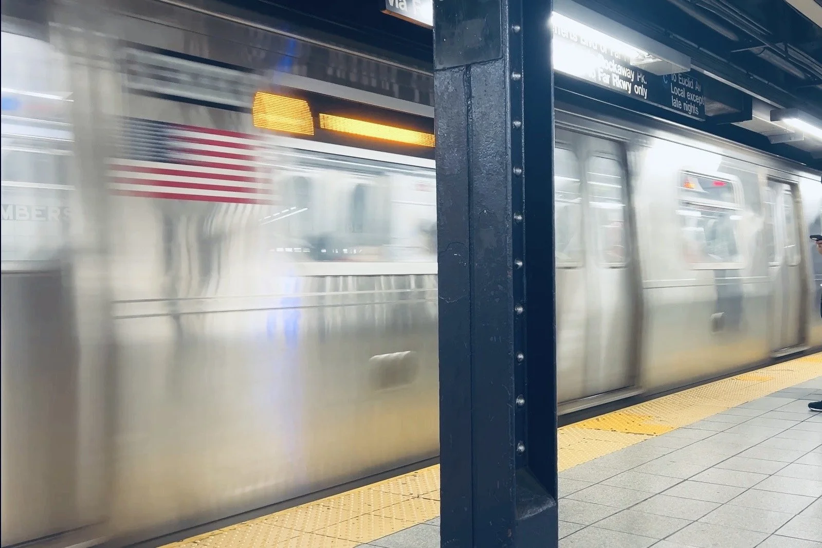 Motion blur shot of a New York City subway train pulling into an underground station past a dark metal pillar and a bright yellow safety platform strip.