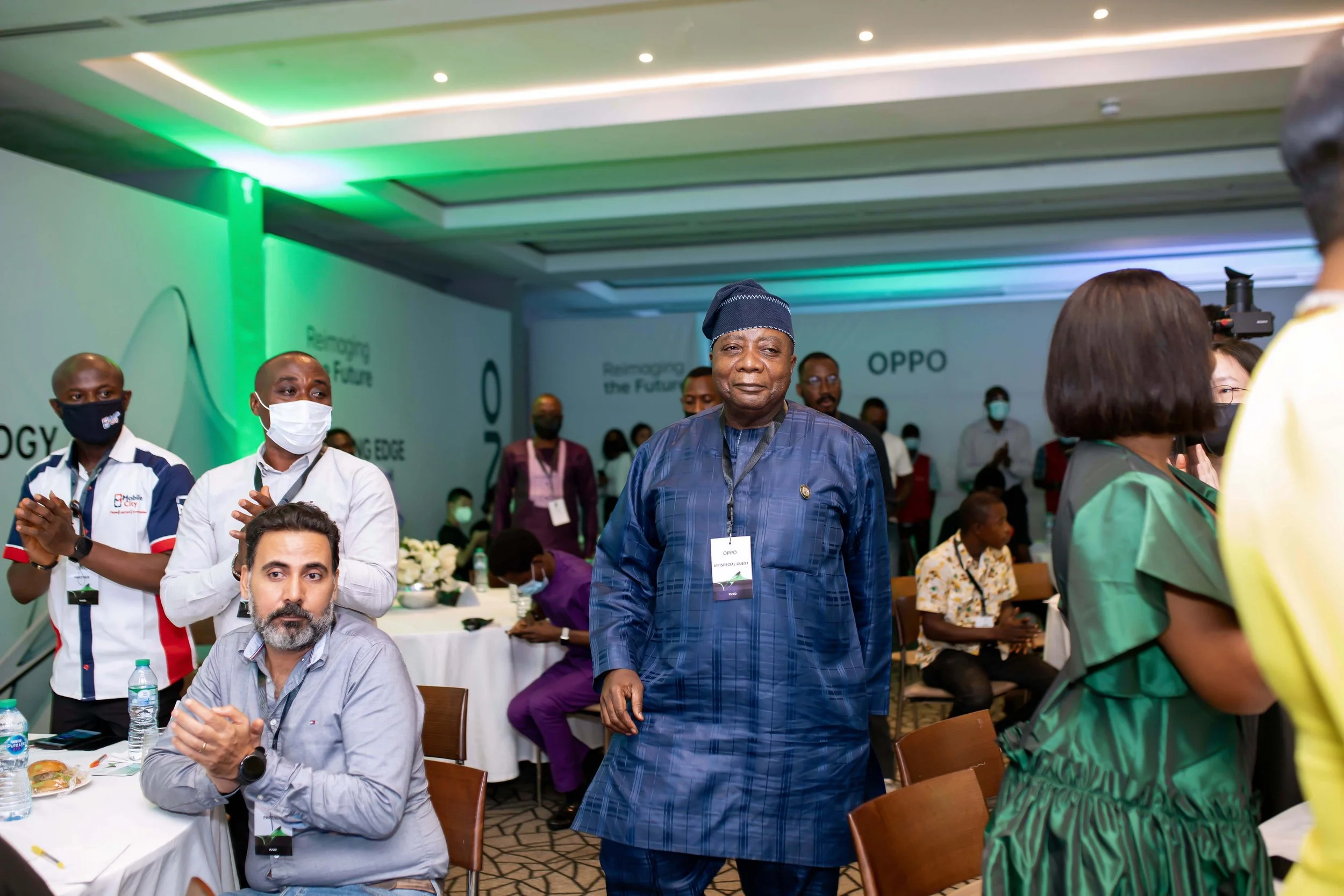 A group of people attending an OPPO event, some wearing masks. An older man in traditional African attire stands in the center. The background features a green-lit wall with the words 'Forging the Future' and 'OPPO' visible.