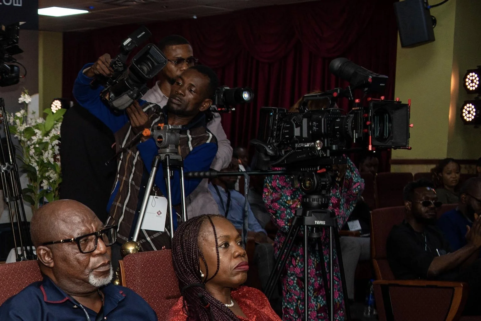People seated in an auditorium, with a man and woman in the foreground and several camera operators filming from behind.