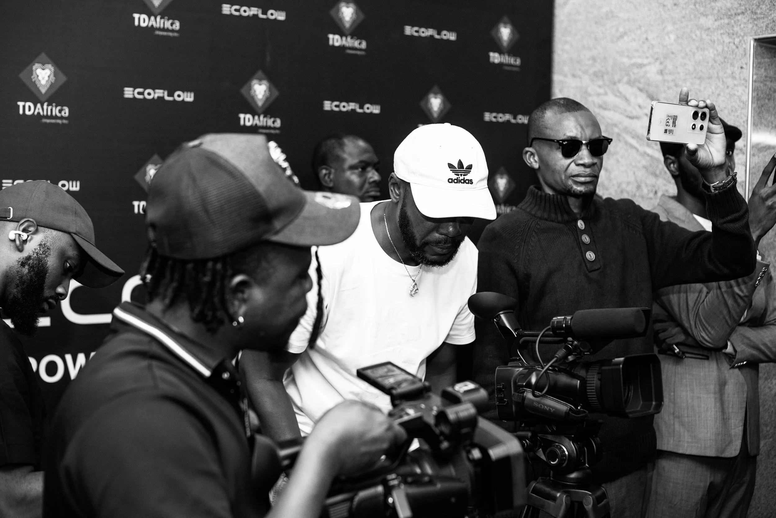 Sane House Media camera operators filming at an EcoFlow event, with a black backdrop titled TDAfrica and EcoFlow behind them. One man is taking a selfie or recording with his phone.