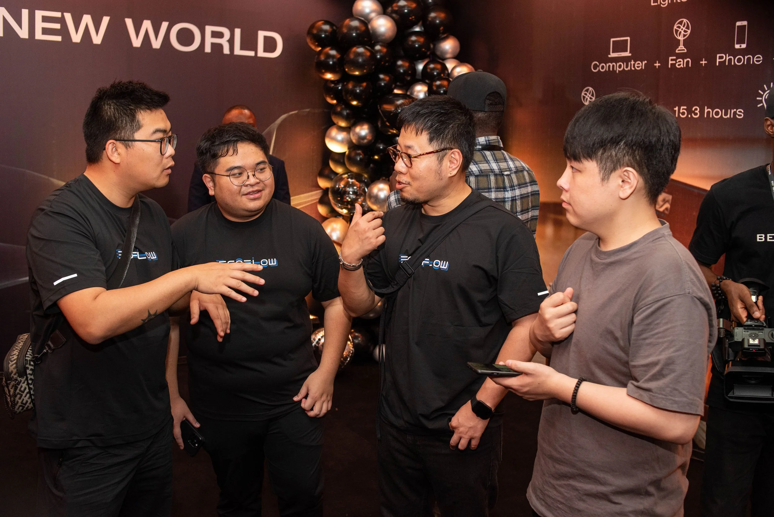 A group of five young men standing together, engaged in conversation at an indoor event with a black wall background and balloons. One is holding a smartphone, and another has a camera. The background features words like 'NEW WORLD' and icons of a co