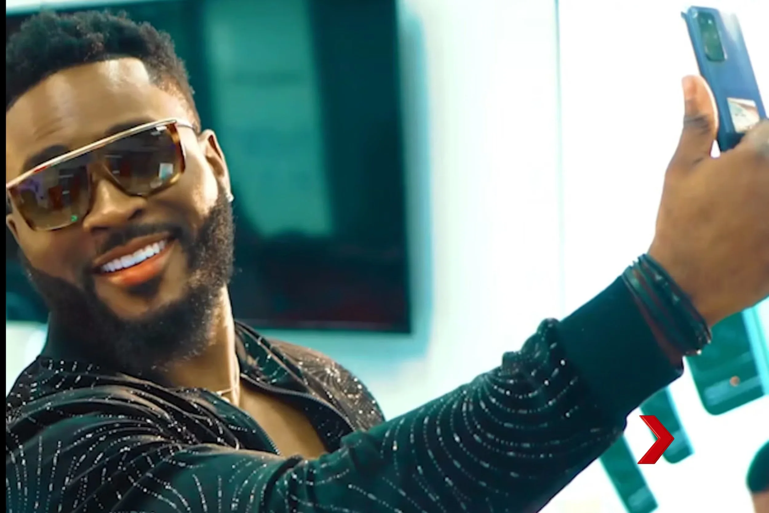 Man smiling wearing sunglasses taking a selfie in front of a bright background.