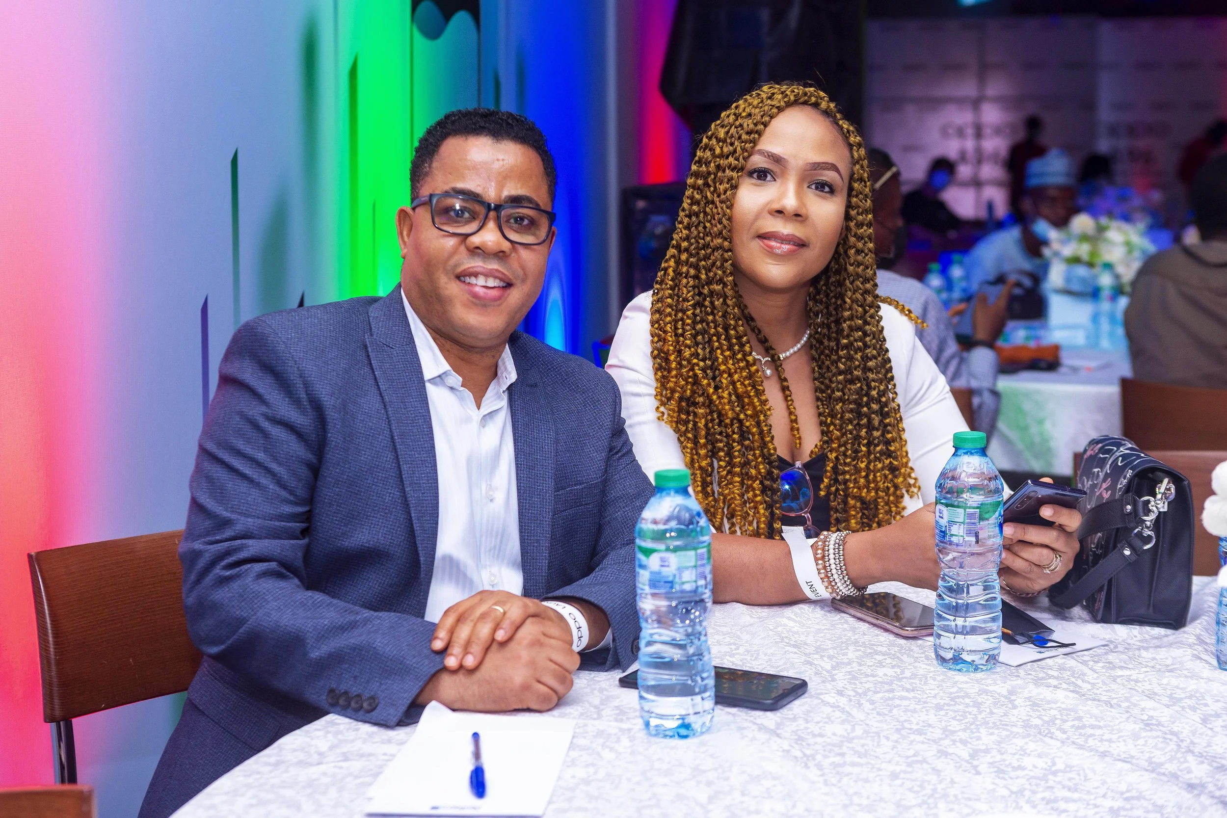 Two people, a man wearing glasses and a woman with braided hair, sitting at a table with water bottles, a phone, and a bag in front of them during an indoor event with colorful lighting and other attendees in the background.