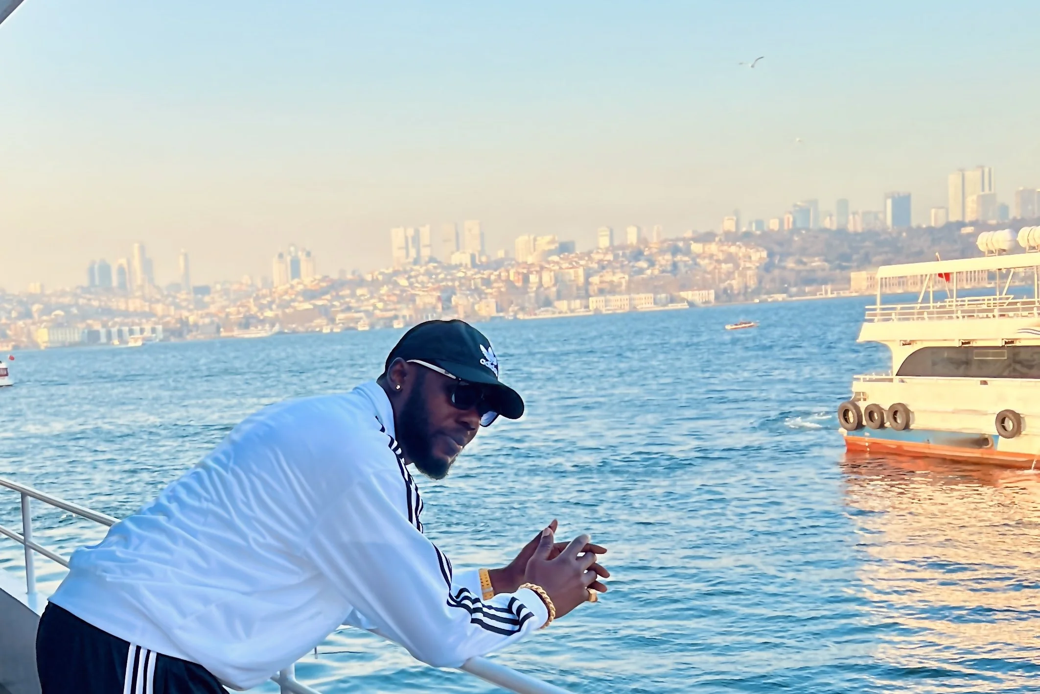 A man in a white Adidas track jacket and black cap leans over the railing on the open deck of a passenger ferry crossing the Bosphorus from Europe to Asia, with Istanbul’s European shoreline visible in the background.
