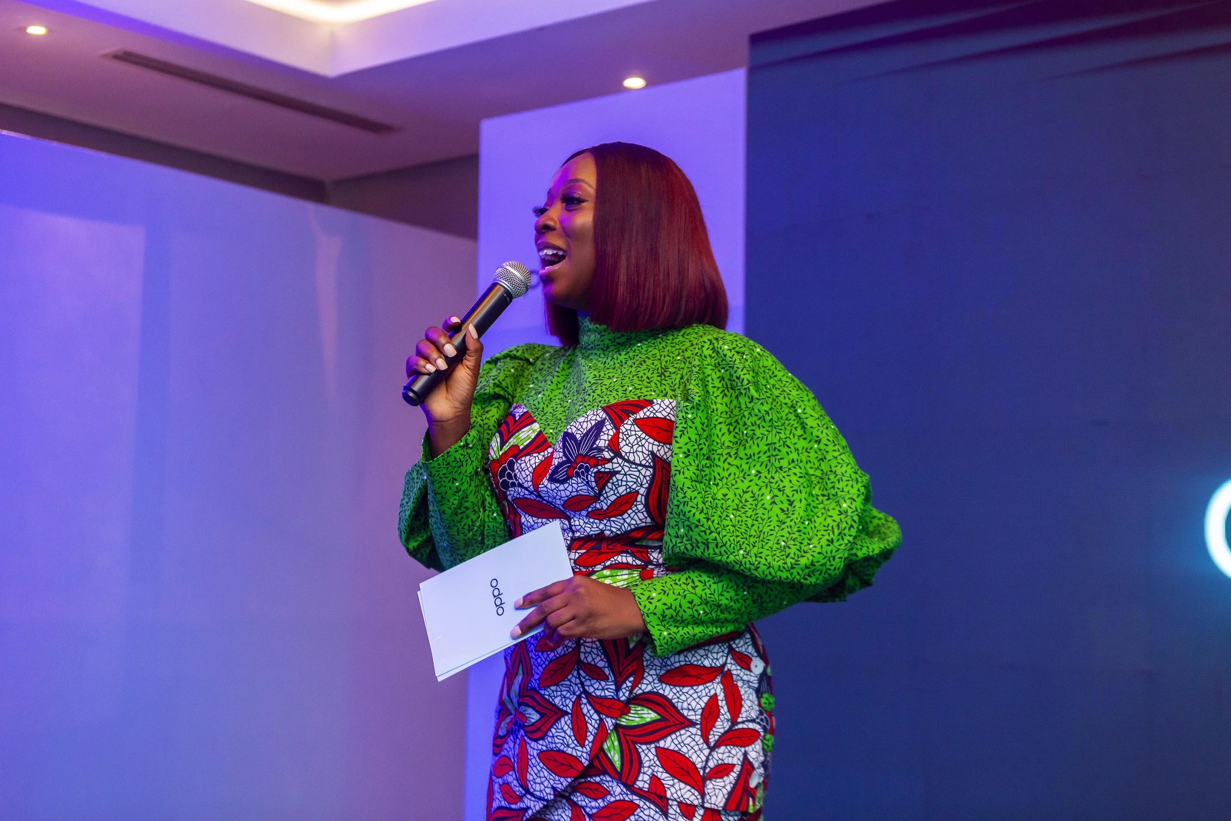 A woman with red hair wearing a vibrant green and red patterned traditional African dress, holding a microphone and speaking at an indoor event with purple lighting.