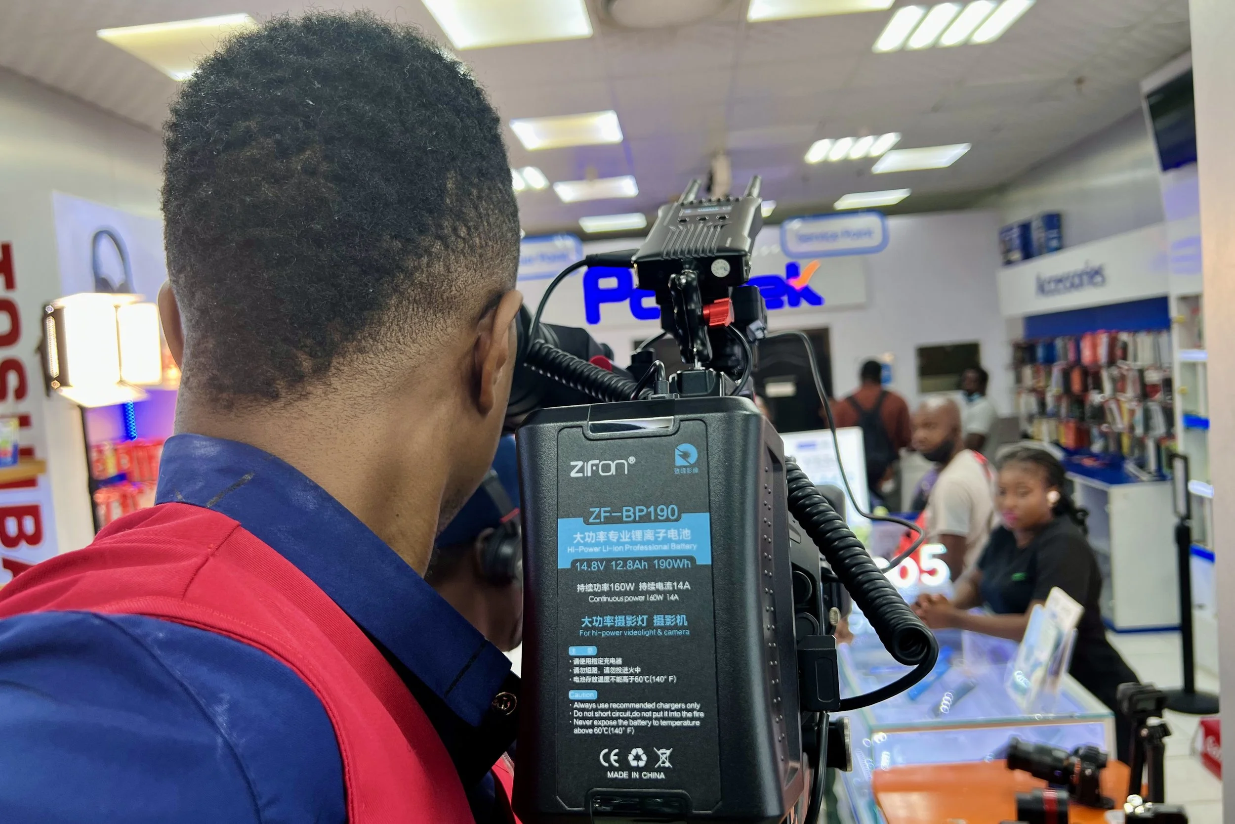 A man operating a professional camera in a retail store, with shelves and customers in the background.