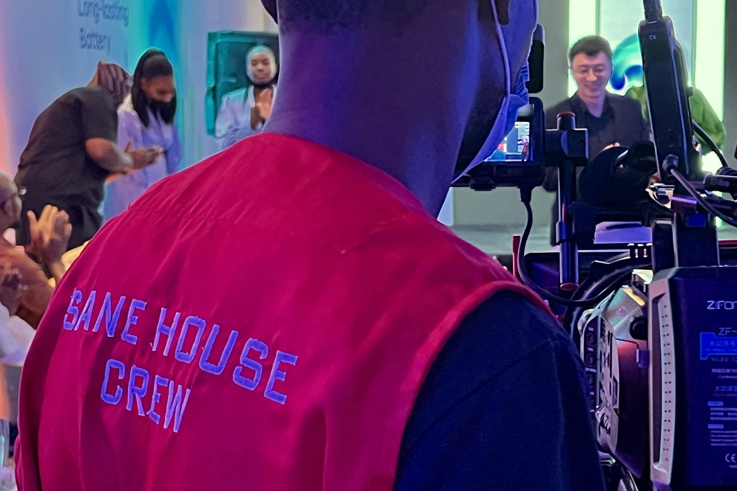 Person wearing a red shirt with partially visible white and purple text, standing in front of a camera setup capturing several people, including a woman in a white coat and a man with glasses smiling.
