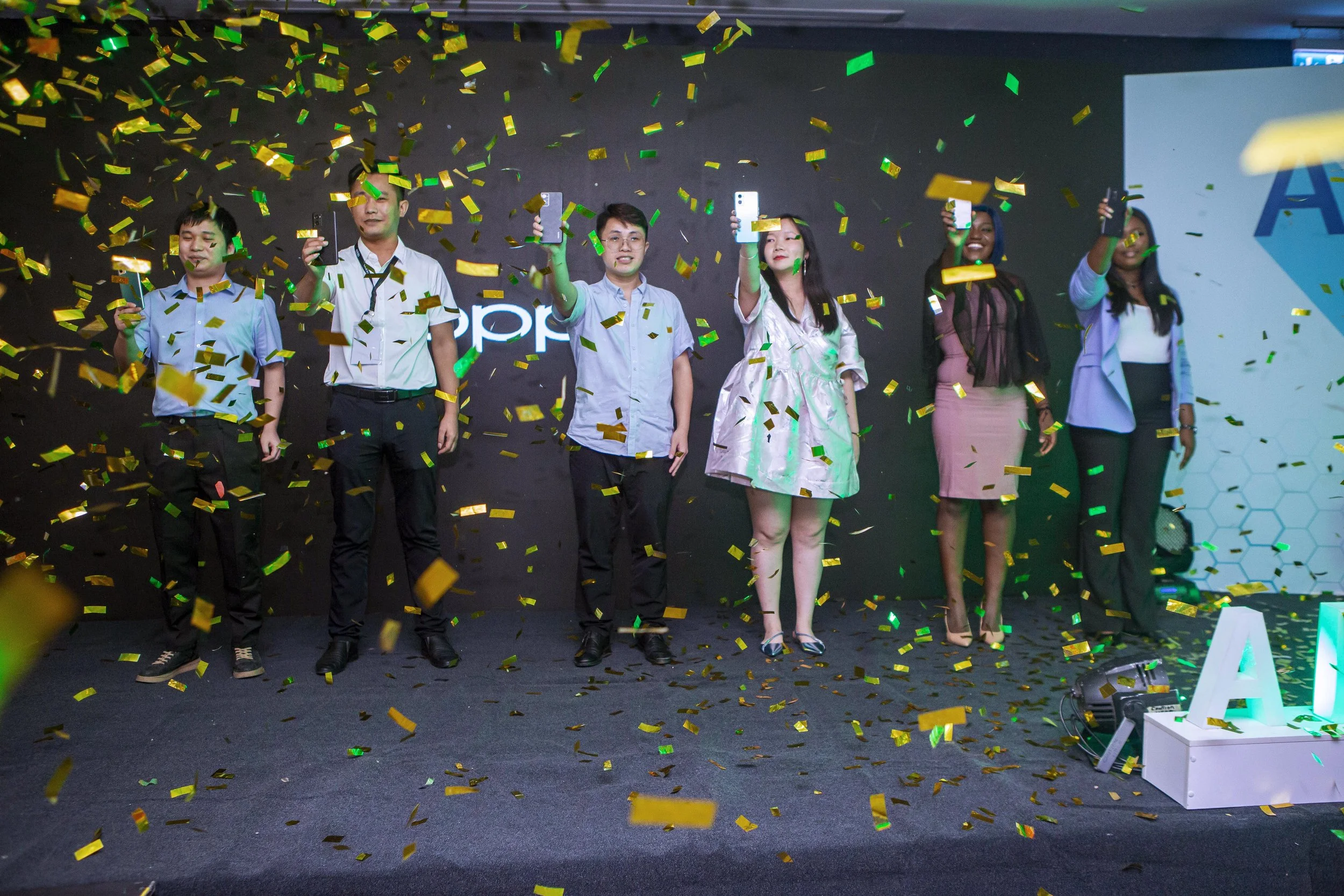 Six OPPO staff on a stage surrounded by falling gold and green confetti, taking photos or videos with their phones. 