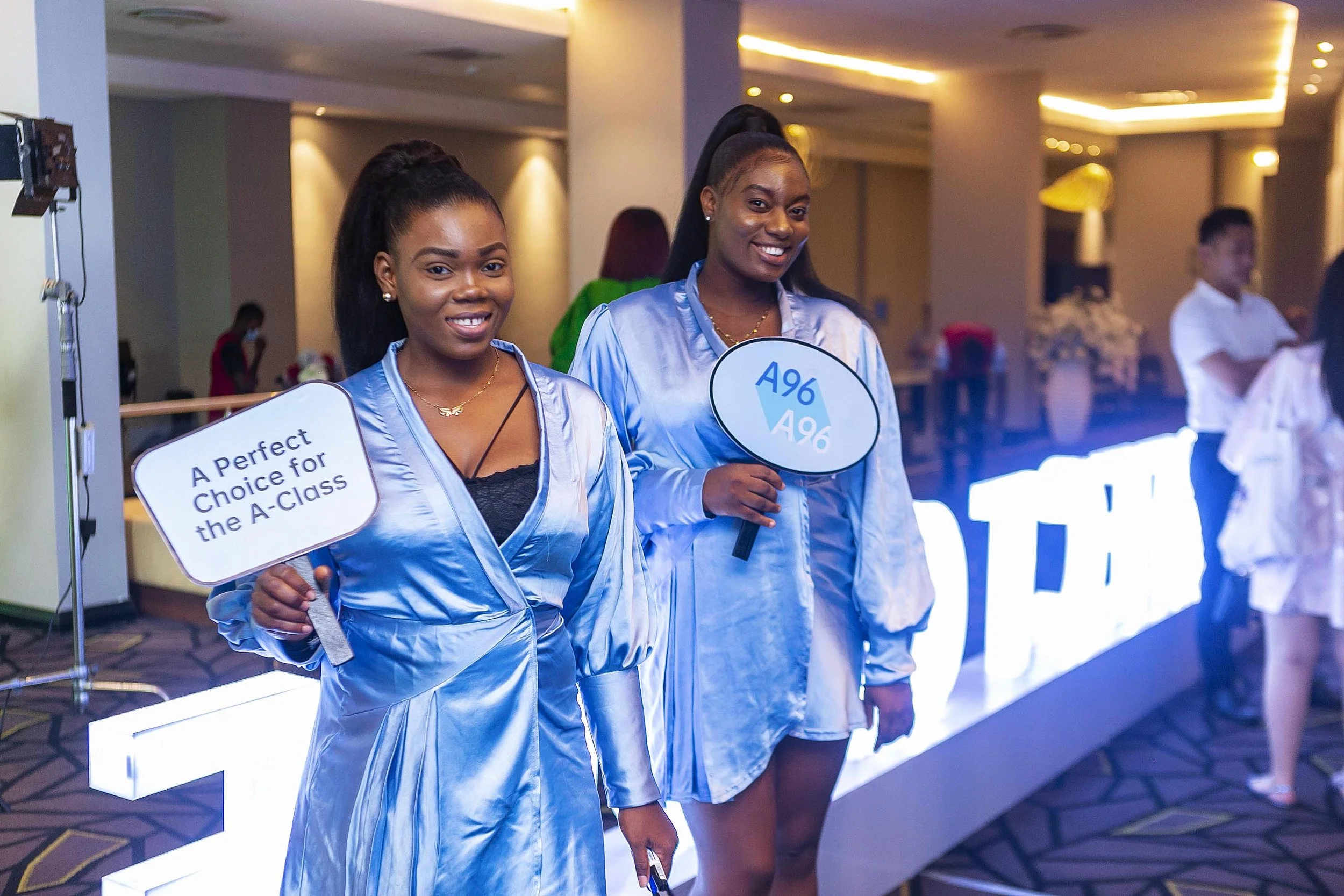 Two women with dark hair in high ponytails, wearing shiny light blue satin robes, standing indoors at an event. One woman is holding a sign that says "A Perfect Choice for the A-Class," and the other has a round sign with "A96" written on it. They ar