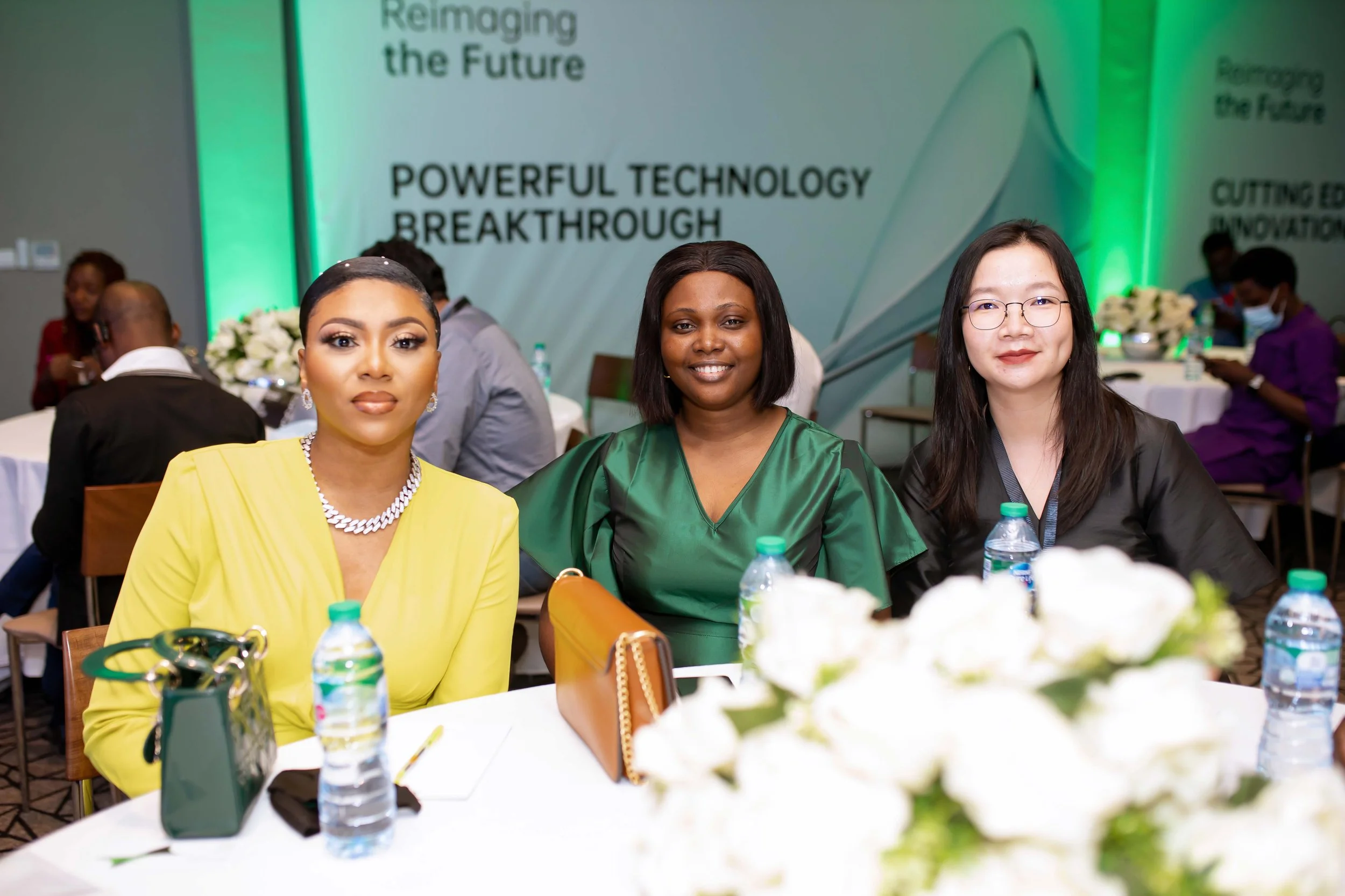 Three women sitting at a conference table, smiling at an OPPO event. A large banner behind them reads 'Reimagining the Future. Powerful Technology Breakthrough.