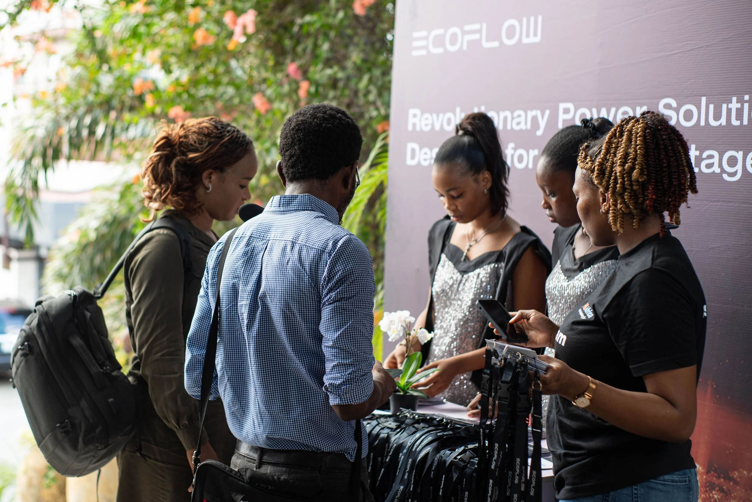 Group of young people at an event near a booth with a large purple sign that reads 'ECOFLOW' and 'Revolutionary Power Solutions.' They are engaged in conversation, some holding brochures or electronic devices, with lush greenery in the background.