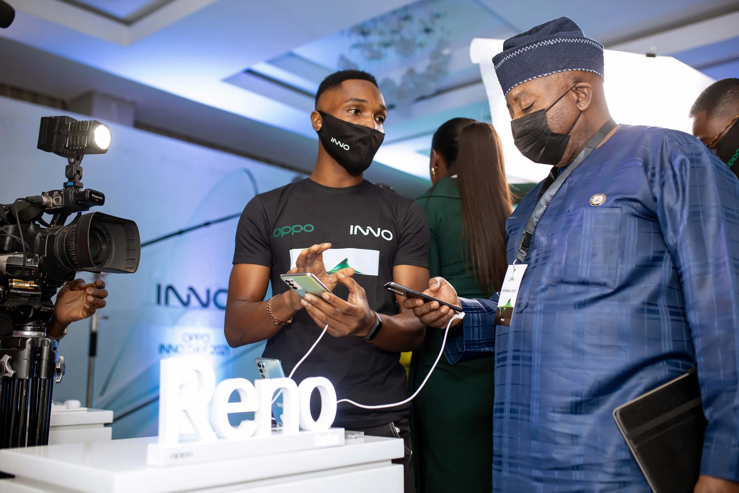 Two men wearing masks and business attire are engaged in conversation at an event, with a professional camera and a white display with the word 'REVO' on it in the background.